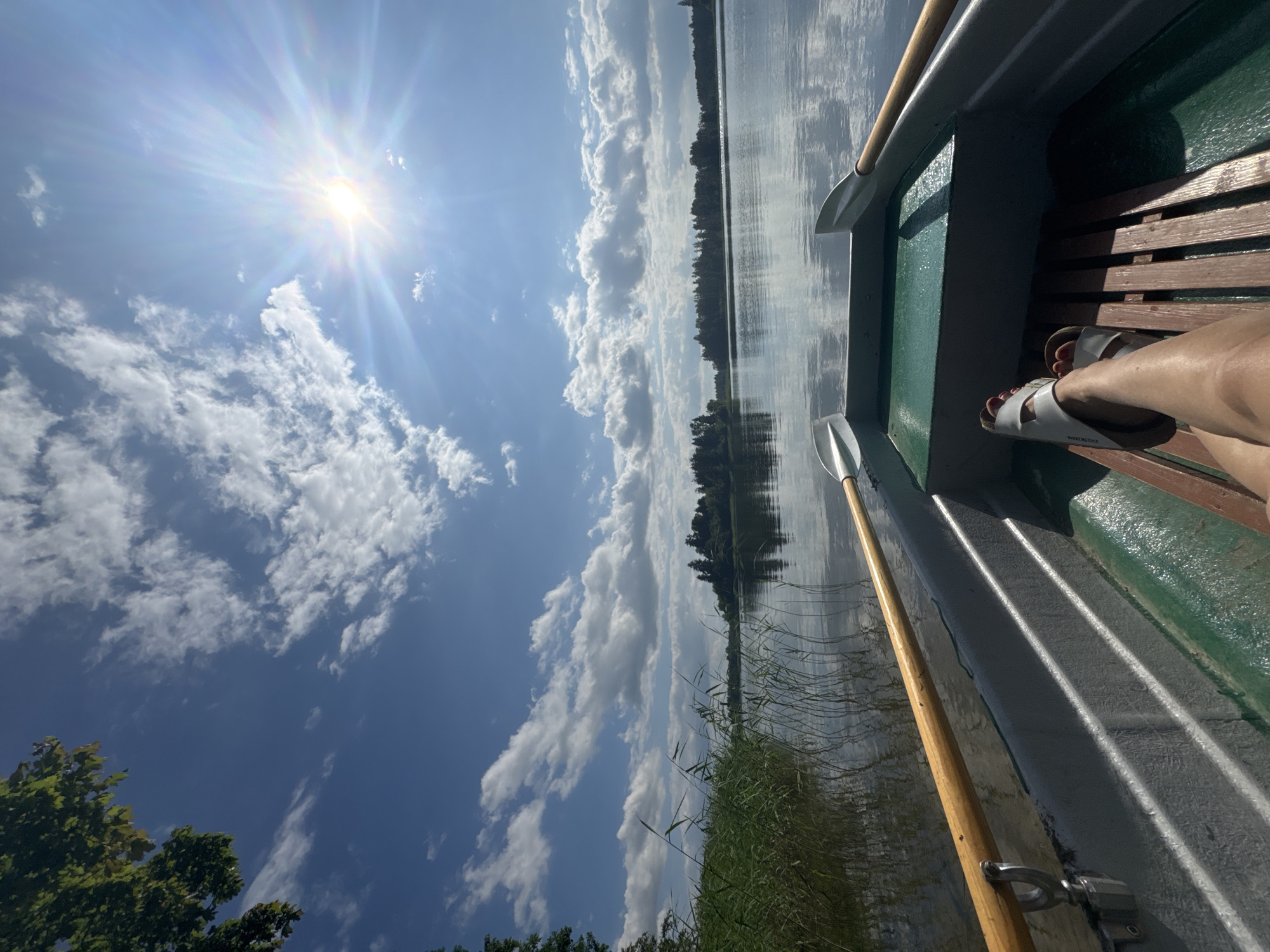 The image depicts a serene scene from a boat on a lake in Lendži, Latvia. The perspective is from inside the boat, looking out towards the water and the sky.
The **subjects** include the interior of a boat with green gunwales and wooden benches, a person's legs and feet wearing white sandals, and two oars. The **scene** is a tranquil lake surrounded by lush green trees and reeds along the shore. The **setting** is clearly outdoors, on the water, under a bright sky.
The **activity** suggested is leisurely boating or rowing, implied by the presence of oars and the seated posture of the person.
**Notable details** include the brilliant sunshine, indicating it is a bright, sunny day, likely daytime. The sky is a vibrant blue, dotted with fluffy white clouds, suggesting fair **weather**. The reflection of the sky and the surrounding trees on the calm water adds to the peaceful atmosphere. The sun's rays are prominently featured, creating a lens flare effect. The presence of a person suggests a moment of relaxation and enjoyment.
There is **visible text** on the sandal worn by the person, which appears to read "BIRKENSTOCK".