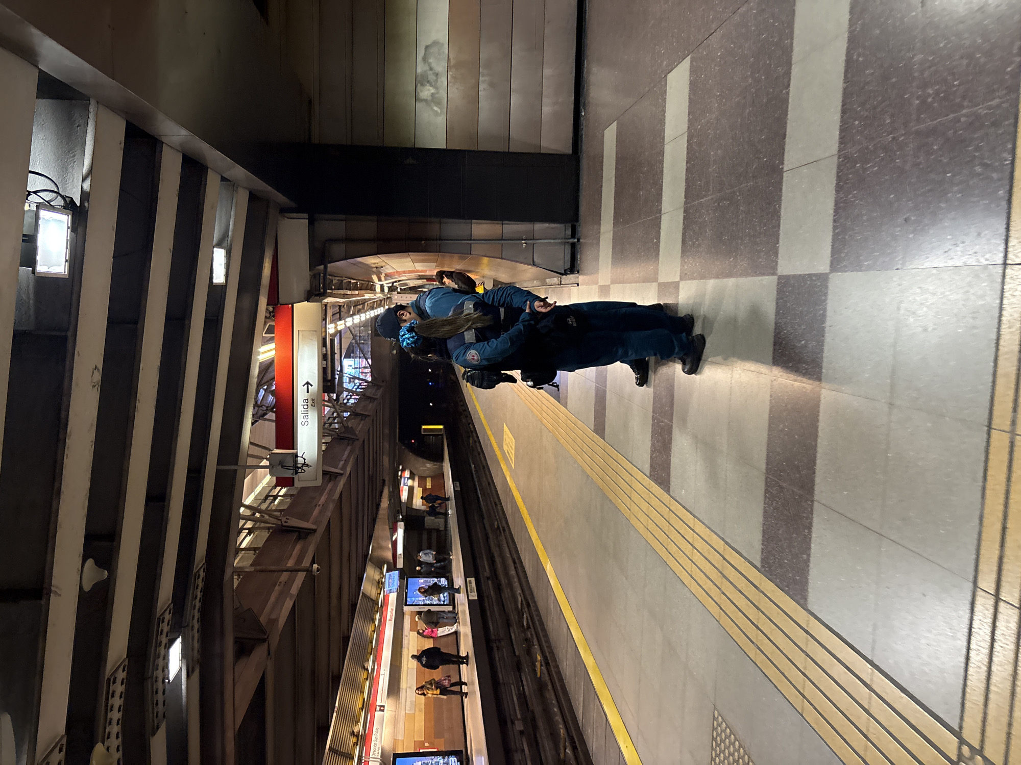 The image depicts a subway platform in Santiago, Chile, likely part of the Metro de Santiago system, given the presence of red and white signage with Spanish text and the characteristic platform design. Two uniformed individuals, possibly security personnel or transit staff, are standing on the platform facing away from the camera. They are wearing dark blue uniforms with patches and appear to be conversing or standing guard. One individual has a blue ribbon in their hair. Other passengers are visible further down the platform, standing near a train. The platform itself is clean, with tactile paving for the visually impaired running along the edge. Overhead, the distinctive architecture of the subway station is visible, with a suspended ceiling structure and lighting fixtures. The overall atmosphere is that of an operational public transport hub during a period of activity.