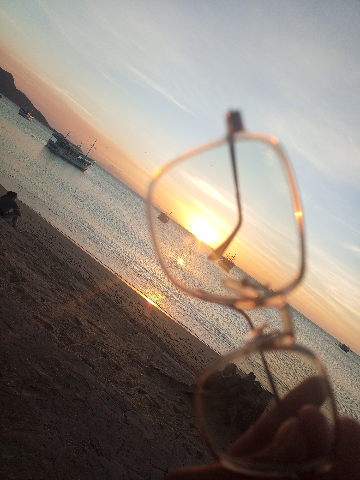 The image displays a coastal sunset scene in Juan Griego, Venezuela, captured with a diagonal horizon line sloping upwards from left to right. In the immediate foreground, a human hand holds a pair of eyeglasses with light-colored frames, which are partially blurred but central to the composition. Bright sunlight reflects prominently off the lenses. The ground in the lower left foreground consists of dark, textured sand.
In the midground, a sandy beach extends towards the left, where a single individual is seated on the sand near the water's edge, facing the calm body of water. This individual is silhouetted and wears dark clothing. Several boats are moored in the water; a larger, white-hulled vessel with a cabin and mast is positioned to the left of center, while another smaller boat is visible further right, directly within the path of the sun's reflection. The water itself is calm, showing a shimmering, golden-orange reflection of the setting sun that extends from the horizon towards the beach.
The background features the sun low on the horizon, emitting intense orange and yellow light. The sky above transitions from orange near the sun to a pale blue further upwards and to the right. A dark, mountainous landmass is silhouetted against the sky on the far left horizon.