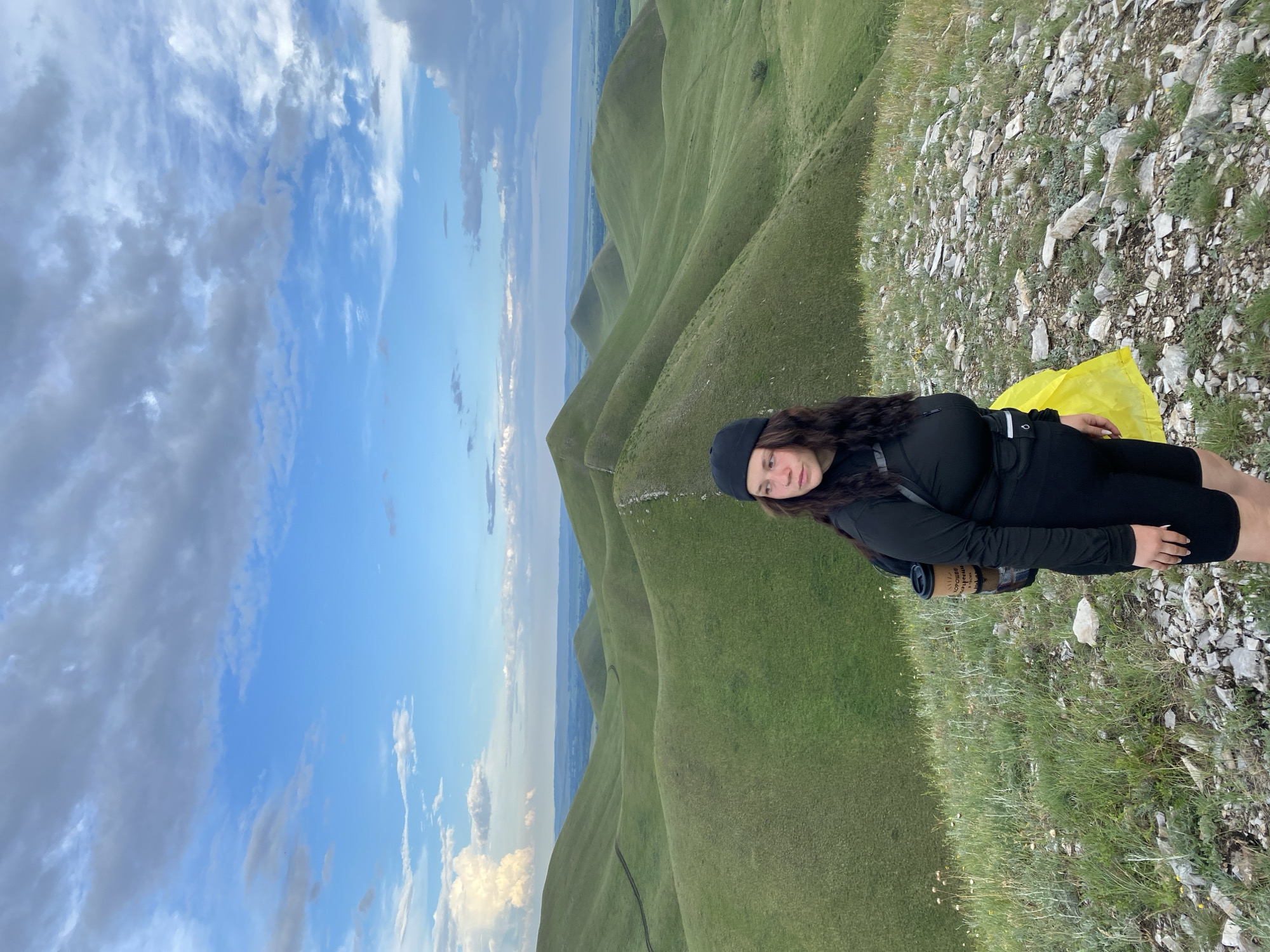 A young woman wearing a black beanie, black long-sleeved shirt, and black shorts stands on a rocky, grassy hillside. She has a backpack on her shoulders with a coffee cup attached to it. A bright yellow object, possibly a bag, is tucked behind her right leg. The background consists of rolling green hills under a cloudy blue sky. The location is described as being near Verkhneozernoe, Russia.