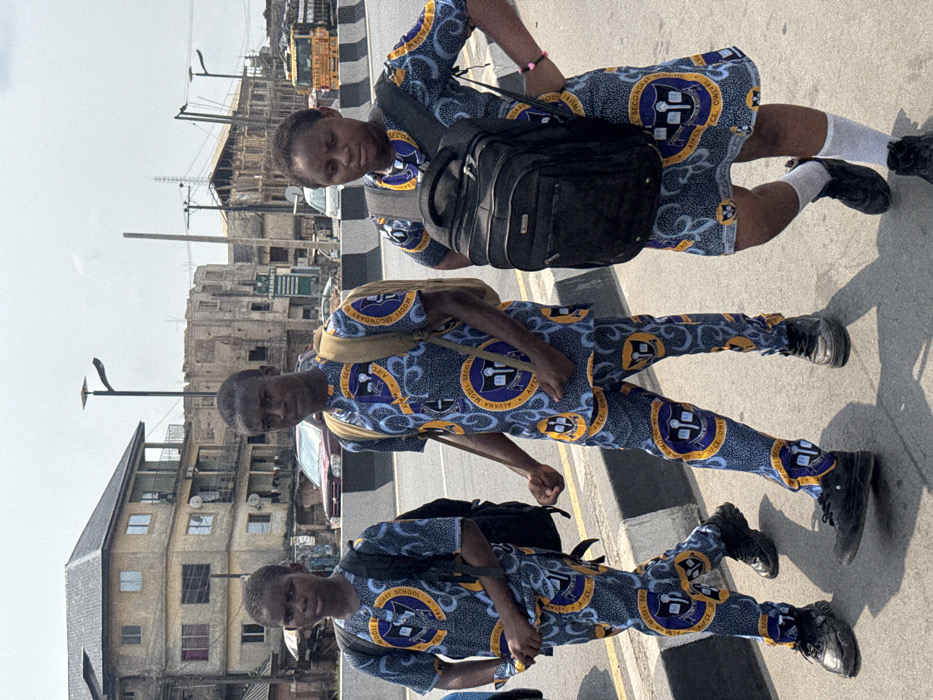The image depicts three individuals, two males and one female, appearing to be teenagers, walking along the shoulder of a paved road in Ogbaku, Nigeria, under a bright, hazy sky. The urban environment features multi-story buildings in the background.
All three individuals are wearing identical school uniforms, consisting of blue, yellow, and white patterned fabric with a circular emblem bearing the inscription "AIYANA MODEL SECONDARY SCHOOL, AIFCO, OWERRI".
The leftmost individual, a male, wears a short-sleeved top and matching trousers. He carries a black backpack on his left shoulder and wears dark athletic shoes. His gaze is directed forward, slightly towards the viewer.
The central individual, a male, also wears a short-sleeved top and matching trousers. He carries a light-colored backpack on both shoulders and wears dark athletic shoes. His gaze is directed forward, slightly towards the viewer.
The rightmost individual, a female, wears a short-sleeved dress. She carries a black backpack on both shoulders, wears white ankle socks and dark athletic shoes, and has a pink bracelet on her right wrist. Her gaze is directed downwards.
The individuals are walking towards the right side of the frame on a paved area separated from the main road by a black and white striped curb. Vehicular traffic is visible on the main road, including a dark red sedan and a yellow truck with "DANGOTE CEMENT" lettering. In the background, several buildings are present, including a light-colored, multi-story structure with balconies and a partially constructed building with exposed framework. A green sign with "PHARMACY" text is visible on one building. Streetlights with solar panels and utility poles are also visible in the scene. No direct interaction between the individuals is observed.