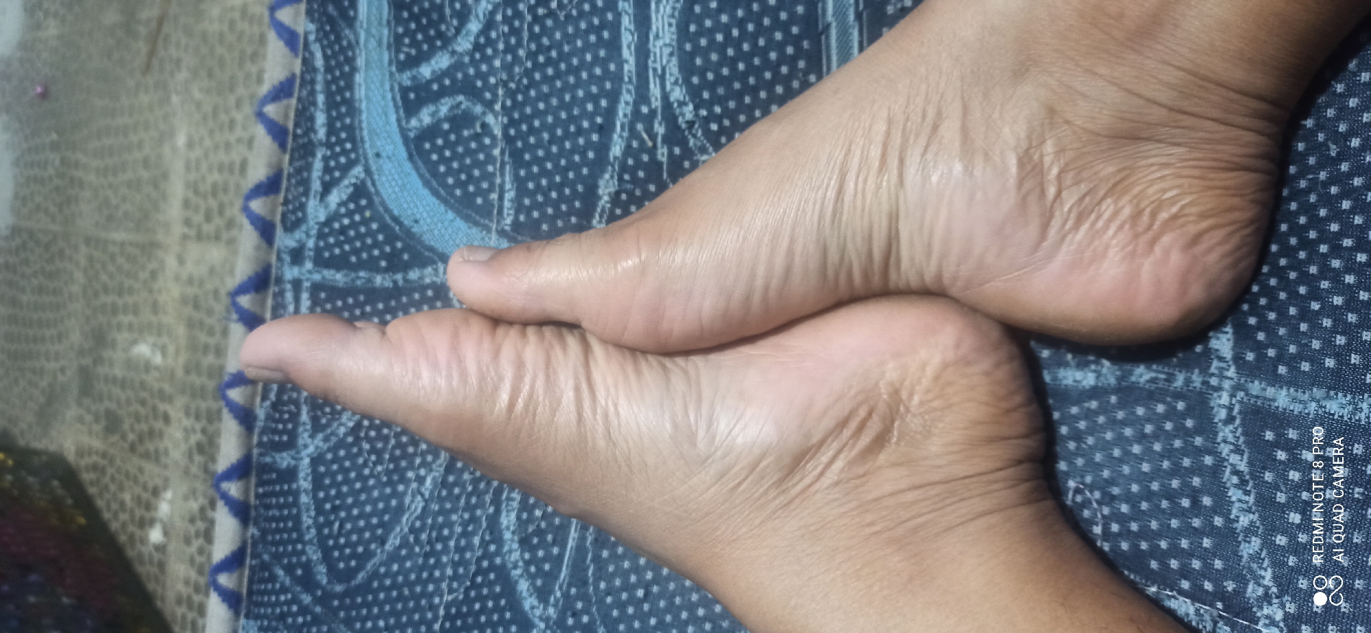 The image shows a close-up view of two bare feet resting on a textured blue surface with a patterned design. The feet are positioned with the soles facing upwards and the toes pointing towards the top of the frame. The skin on the feet appears to have visible creases and texture. In the background, a blurry object with multiple colors and a dark, granular texture is visible. The overall impression is that of a casual indoor setting. The image is a photograph taken with a mobile device, indicated by the watermark "REDMI NOTE 8 PRO AI QUAD CAMERA" in the lower left corner, which suggests the location is likely within Indonesia, possibly in the area of Sedati Agung.