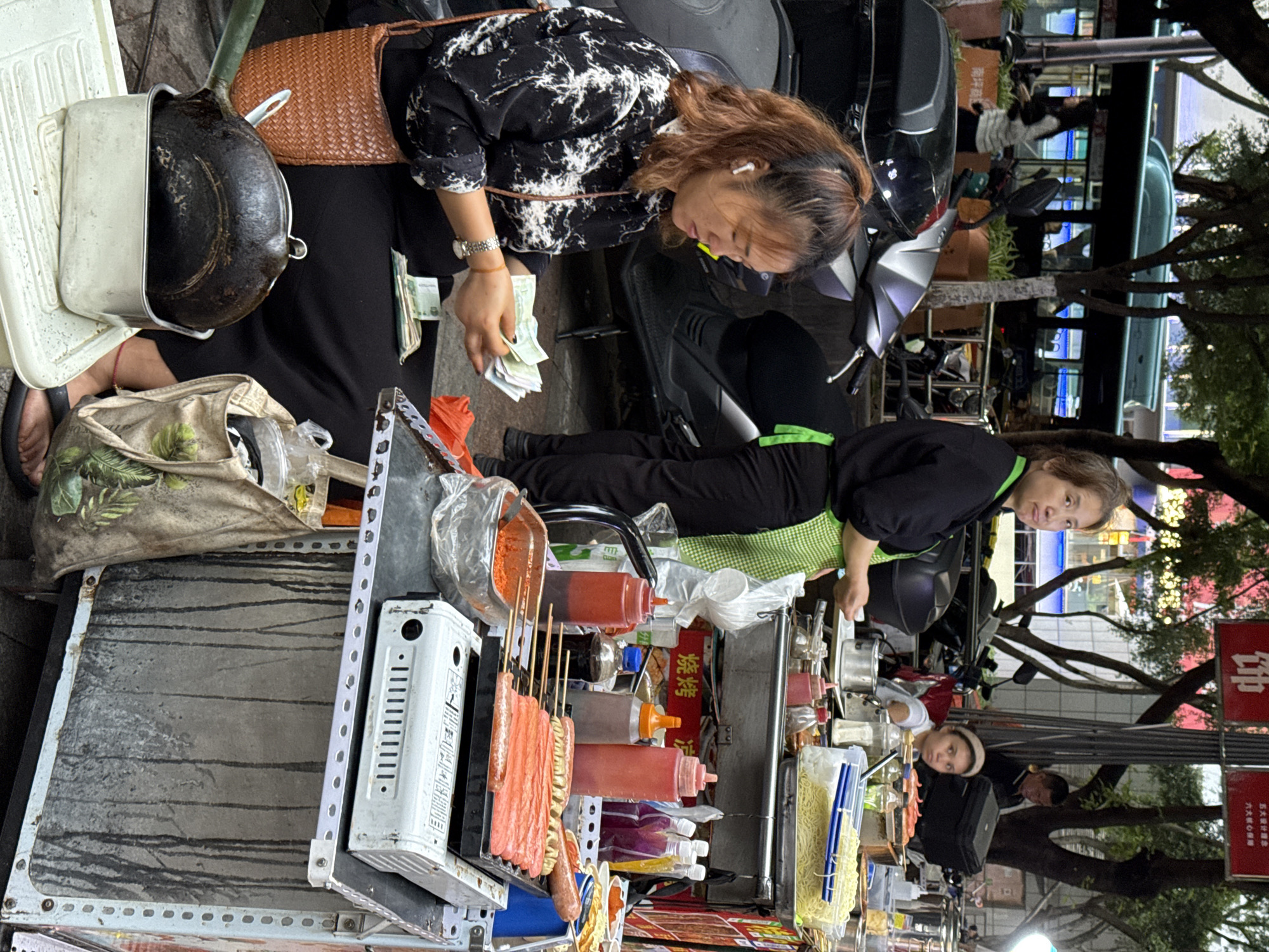 In Nan'an District, China, a woman counts money near a street food cart. The cart is laden with skewers, condiments, a gas stove, and noodles. A second woman, wearing a green apron, stands behind the cart, attending to customers. Passersby are visible in the background.