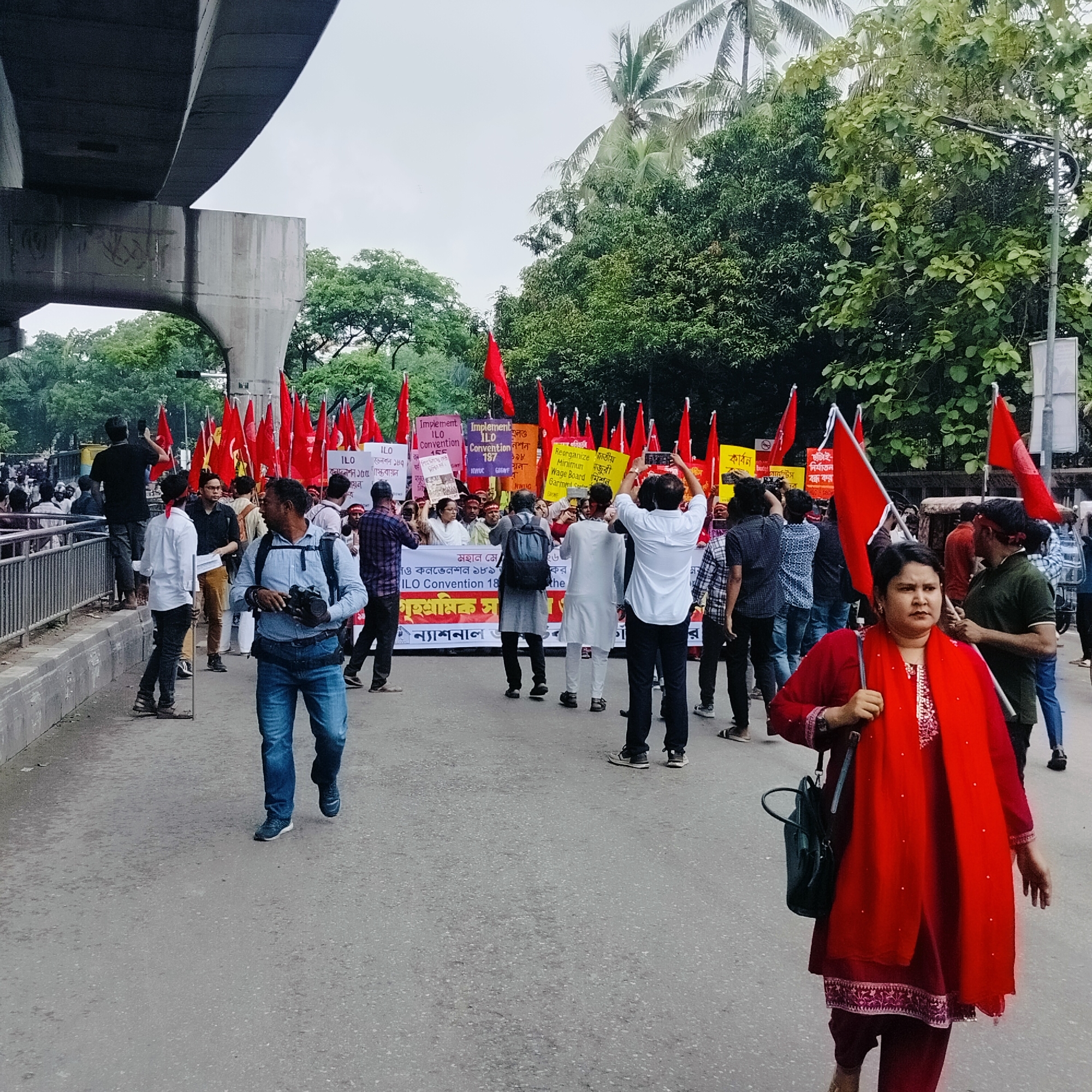 A bustling outdoor scene captures a protest or demonstration under a concrete overpass or elevated road structure, likely in an urban setting. A large group of people, primarily men and some women, are gathered on a street, many holding red flags and various protest signs. Lush green trees and palm trees are visible in the background, suggesting a tropical or temperate climate.
In the foreground, a woman wearing a vibrant red traditional outfit (kurta and scarf) with embroidered details walks towards the right, holding a red flag pole. She carries a dark green shoulder bag and has a serious, determined expression. Beside her, a man walks with a camera in his hand and a backpack on, dressed in a blue shirt and jeans. Other individuals throughout the crowd are seen taking photos or videos with their mobile phones.
The protest signs, some in English and some in Bengali, clearly advocate for labor rights. Visible English text includes "Implement ILO Convention 155," "Implement ILO Convention 187," "ILO Convention 189," and "Reorganize Minimum Wage Board Garment Sector." Bengali text on the signs also mentions "জাতীয় মজুরী কমিশন" (National Wage Commission), "গৃহশ্রমিক" (domestic worker), "ছাঁটাই-নির্যাতন বন্ধ কর" (Stop layoffs/harassment), and "মহান মে" (Great May). Acronyms like NWUC, GAGNF, and NWIG are also present on some placards. The overall mood appears engaged and purposeful. The sky is overcast, suggesting daytime.