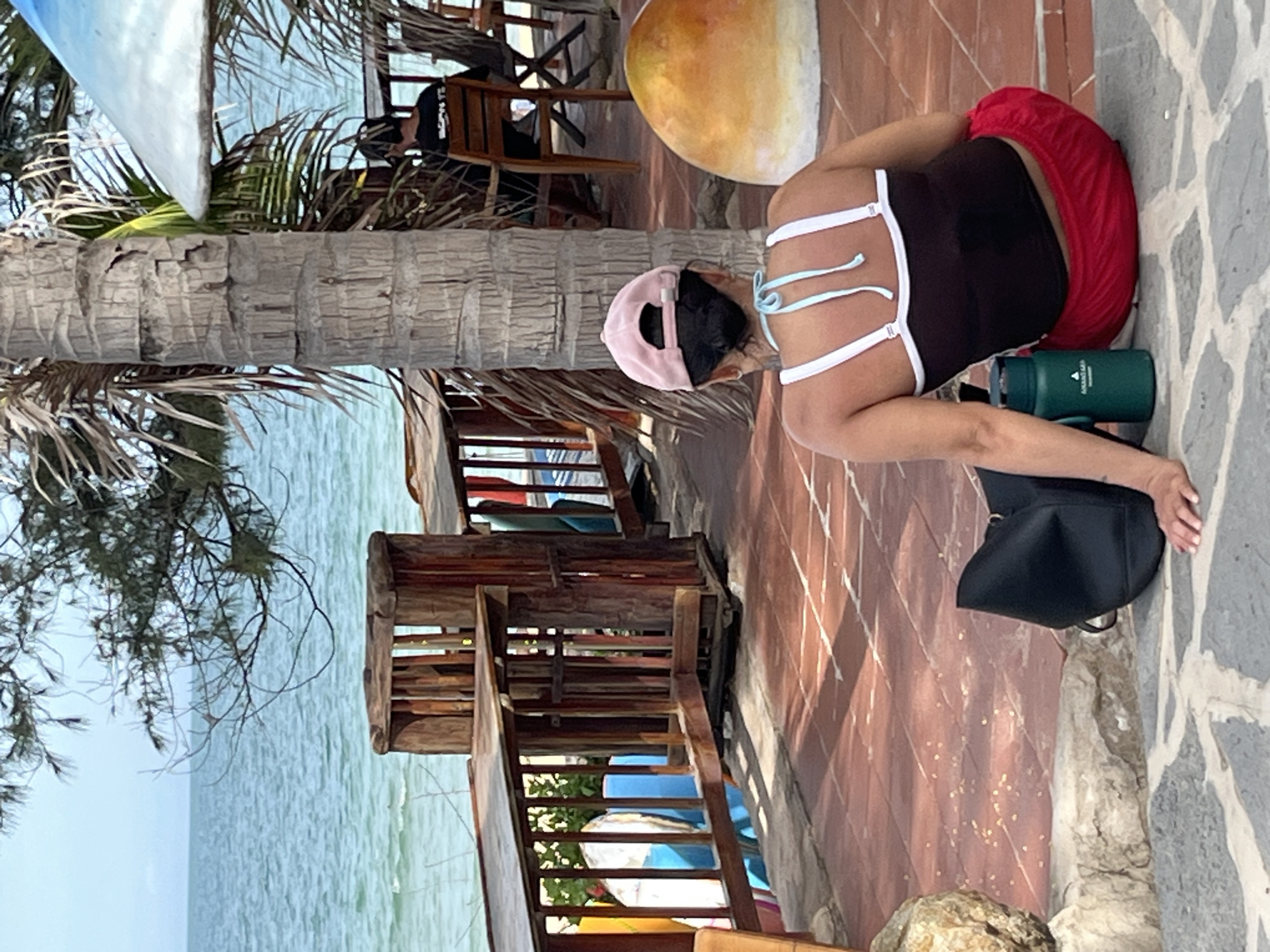 A woman with a pink baseball cap sits facing away from the camera on a tiled patio, near the beach in Phu Thuy Ward, Vietnam. She is wearing a black tank top with white straps and bright red shorts. To her left, a black tote bag rests on the ground next to a green water bottle. The patio is paved with terra cotta tiles and bordered by stone. Beyond the patio, a wooden fence overlooks a calm, blue ocean. Trees with sparse leaves and palm fronds frame the background. In the distance, a person sits at a table under a white umbrella.