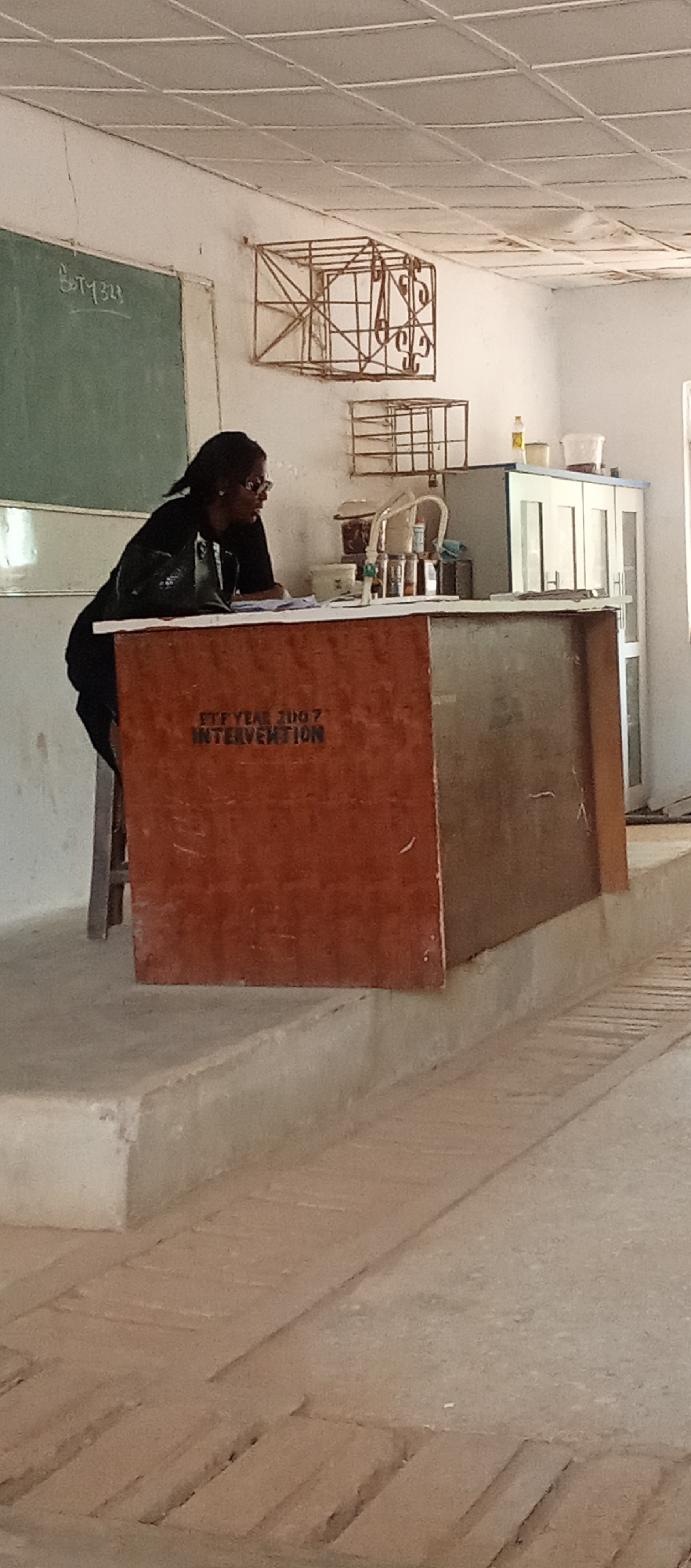 A woman in a dark jacket is seated at a wooden table/counter. The table has the inscription "ETF YEAR 2007 INTERVENTION". The room has a concrete floor with rectangular grooves. In the background, there is a green chalkboard with writing on it, as well as metal cage-like structures affixed to the wall and a cabinet. The location is suggested to be Akwanga, Nigeria. The scene appears to be inside a classroom or laboratory.