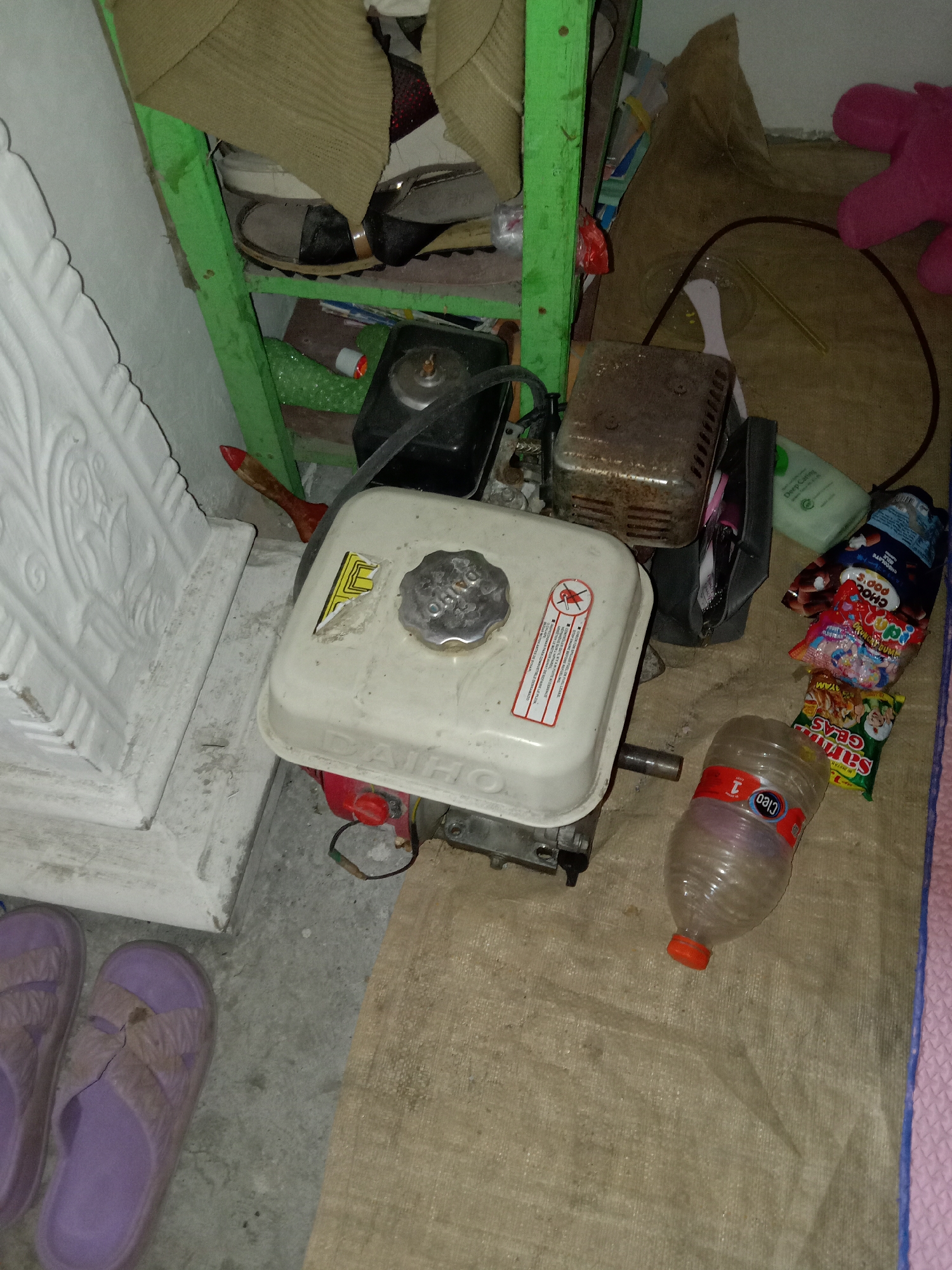 In a dimly lit interior, likely a storage area in Pacing, Indonesia, a small gas-powered engine sits on a tan tarp. The engine is white and red, with a metal cap on top. To its left, a white decorative pillar stands against a wall. Next to the engine, a clear plastic bottle with a red cap is visible, and a variety of snack packages are scattered nearby. Behind the engine, a green shelf unit holds various items, including what appear to be shoes and other miscellaneous objects. A pink stuffed toy is visible in the upper right corner of the frame. The floor is a mix of concrete and tarp. The overall impression is one of a cluttered, utilitarian space.