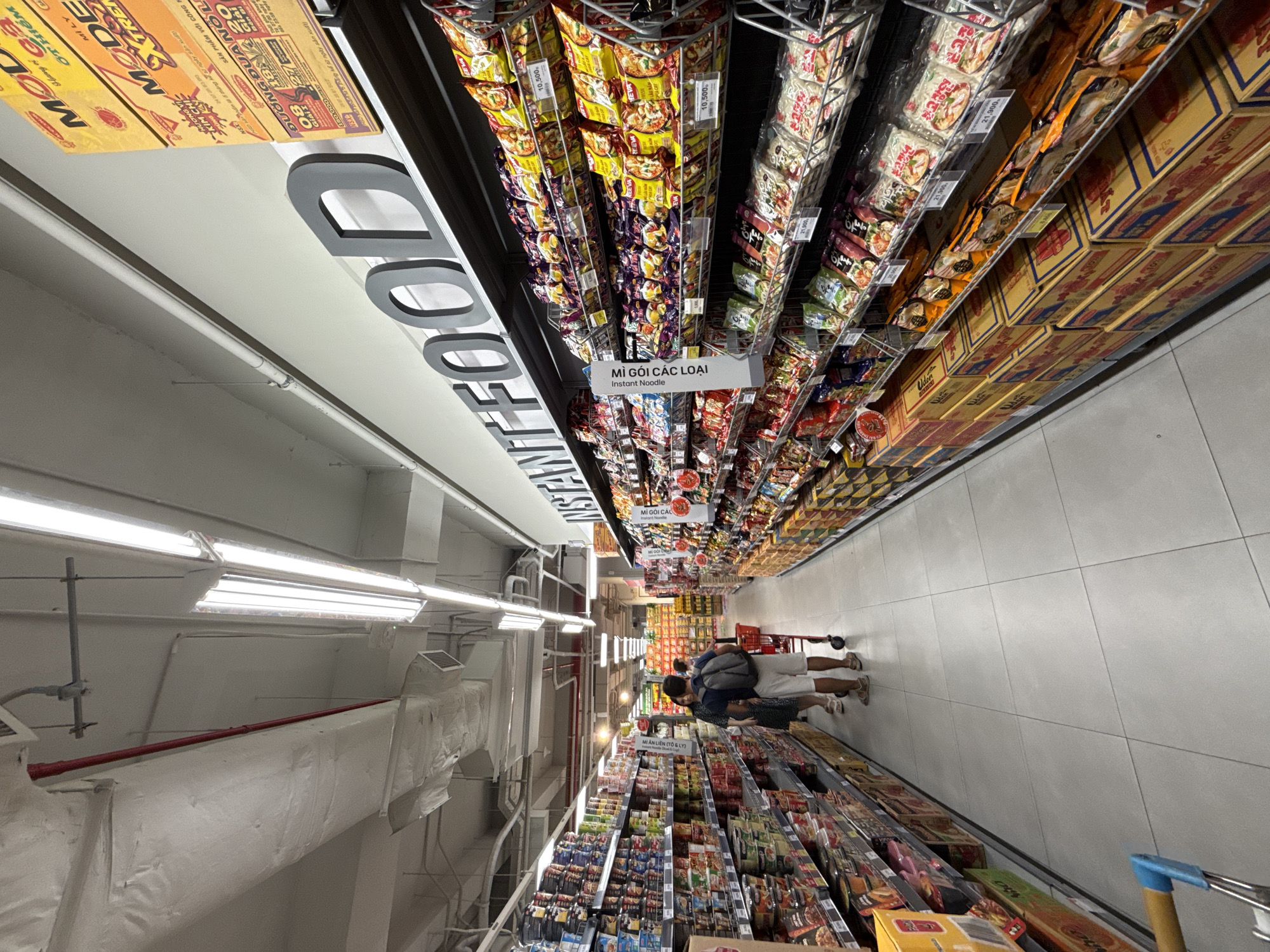 The image shows an aisle in a supermarket in Nha Trang, Vietnam, with a wide selection of instant noodles and other food products displayed on shelves. In the foreground, a shopping cart is partially visible, suggesting active shopping. Two people, a man and a woman, are walking down the aisle, seemingly browsing the products. The man is wearing a backpack, and both are casually dressed. The shelves are fully stocked with various brands and types of instant noodles, with some products featuring prominent signage in Vietnamese. The lighting is bright, typical of a retail environment, and the ceiling has exposed ductwork and lighting fixtures, indicating a modern commercial space. The overall impression is a well-organized and bustling supermarket.