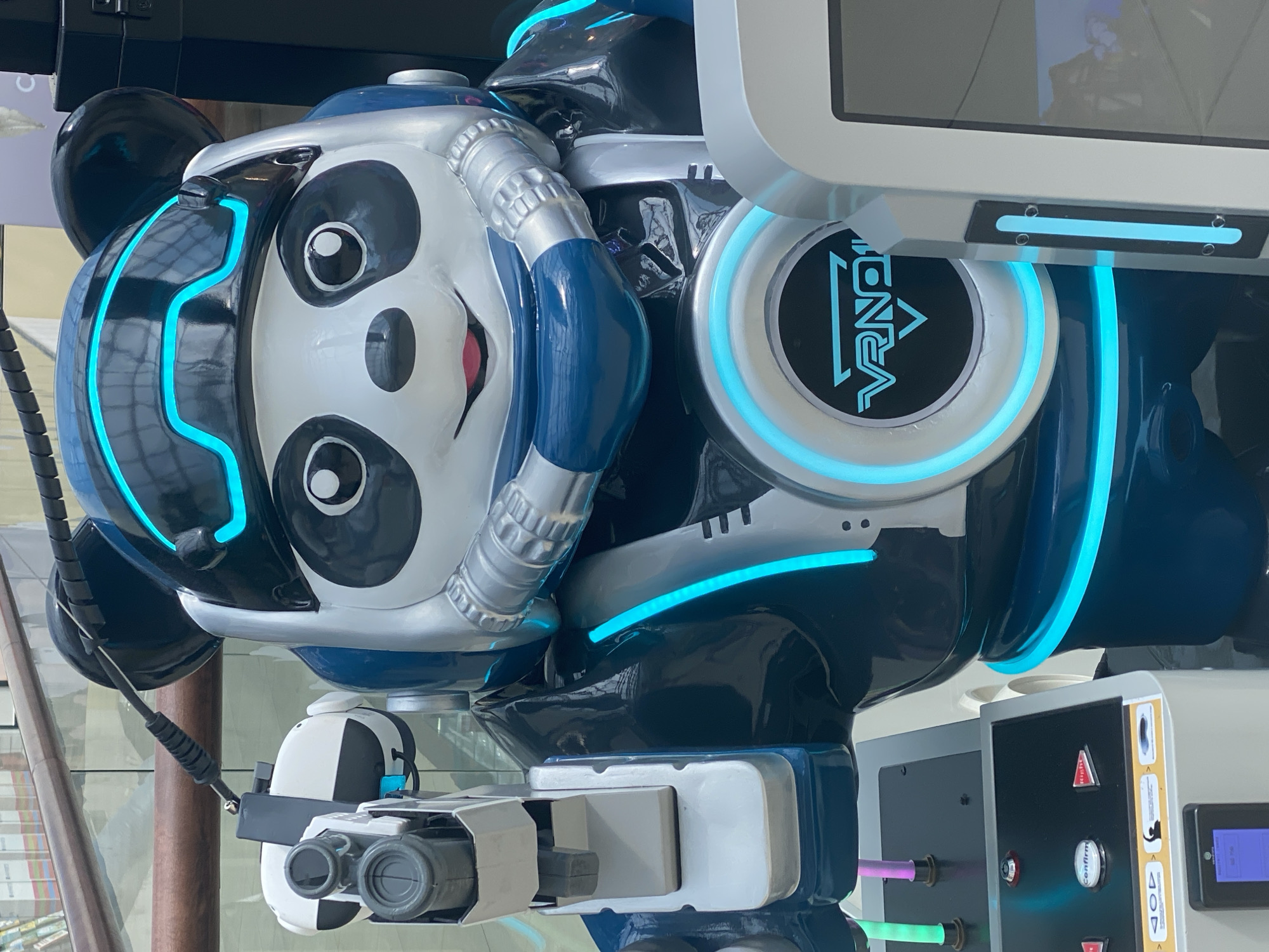 A life-sized, robotic panda character, designed for a virtual reality experience, stands prominently in what appears to be a public indoor space. The robot is primarily white and blue, with black accents around its eyes and ears, resembling a panda. It wears a futuristic helmet with glowing blue trim and has silver, metallic-looking components on its chest and neck, including what resemble air hoses. In its right hand, it holds a gray and white robotic device that looks like a gun or controller. The robot is positioned in front of a kiosk or display unit with a screen and various buttons. A circular emblem with blue neon lighting and the text "VRNO" is visible on the robot's chest. The background suggests a modern building interior with wooden railings and glass, possibly a shopping mall or an exhibition hall. There are no people visible in the image. The image is taken from a slightly low angle, looking up at the robot. The location of Novokuznetsk, Russia, is not discernible from the visual information in the image.