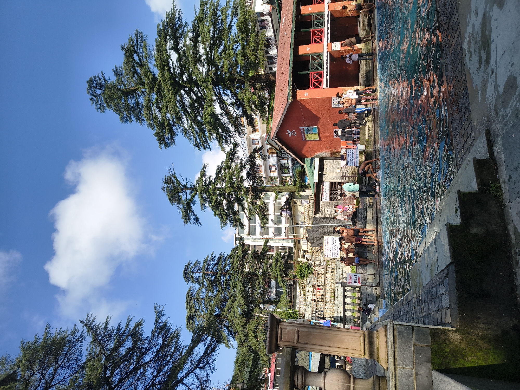 In the city of Bhagsu, India, a bustling outdoor swimming pool is captured under a bright blue sky with scattered white clouds. Tall, mature trees with dense green foliage frame the scene, their branches reaching towards the sky. In the background, a multi-story building with white and beige facades rises above the surrounding structures, interspersed with smaller buildings, including a vibrant orange one with a red roof and green accents.
The pool itself is the central focus, filled with clear, turquoise water that reflects the surrounding light and the figures present. The water's surface is dynamic, with ripples and reflections indicating activity. Stone steps and walkways border the pool, some adorned with moss and greenery.
Numerous people are visible around and in the pool, engaged in various activities. Some are standing in shallow water, while others are near the edge, possibly waiting to enter or exiting. A group of young men, some wearing swim trunks, are gathered on the steps leading into the pool. One individual is bent over at the edge of the pool, appearing to be adjusting their swimwear. Further back, near the buildings, a few individuals are standing and conversing. Signs with text in Hindi and English are posted, including one with an arrow pointing to "TOILET." The overall atmosphere suggests a public recreational space on a sunny day.