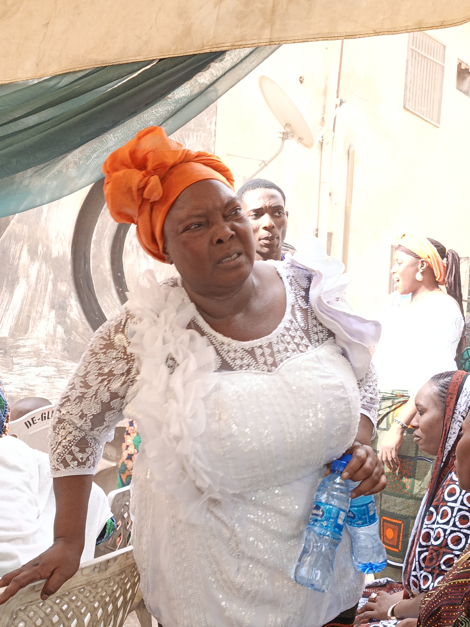 This daytime image captures a gathering in what appears to be Sabon Gari West, Nigeria, likely an outdoor event under a shaded canopy. The central figure is an older woman, prominently featured in the foreground, looking to her right with a focused, perhaps speaking or listening, expression. She is adorned in a vibrant orange gele (headwrap) and a white, long-sleeved dress or top. Her outfit is embellished with intricate lace patterns, sequins, and ruffled fabric detailing across the chest and shoulder. In her left hand, she holds two small plastic water bottles, partially revealing the "CAPRI" brand label.
Behind her, a young man is visible, looking forward with a neutral expression. To the right, further back, another woman is seen with an orange headwrap and braided hair, engaged in conversation. To the far right, a woman in profile wears a patterned headscarf, suggesting traditional attire. Parts of other individuals are also visible, indicating a crowded setting.
The background shows draped fabric in shades of beige and green, suggesting a tent or temporary structure for shade. Beyond this, a light-colored building with a barred window and a white satellite dish is visible. In the lower left, a person is seated, wearing a white band on their head with the partial text "DE-GLI". The bright lighting and visible water bottles suggest a warm climate and a social event.