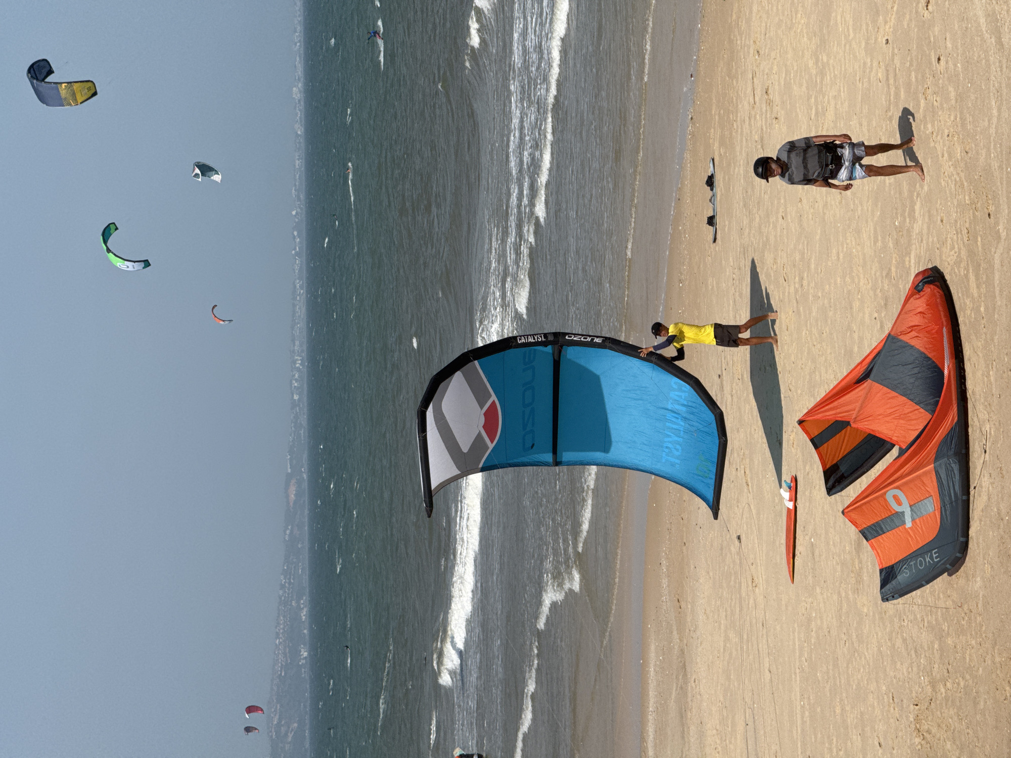 This image depicts a beach scene in Phan Thiet, Vietnam, with several kitesurfers and kites present. In the foreground, two orange and black kites are laid out on the sand. A person in a yellow shirt and black shorts stands near a large blue kite, holding it upright. Further back, a person wearing a striped shirt, shorts, and a helmet is walking on the sand. In the ocean, several kitesurfers are visible, with their kites in the air. The sea is wavy, and there are whitecaps. In the background, a hazy coastline with buildings can be seen. The sky is overcast and light blue.