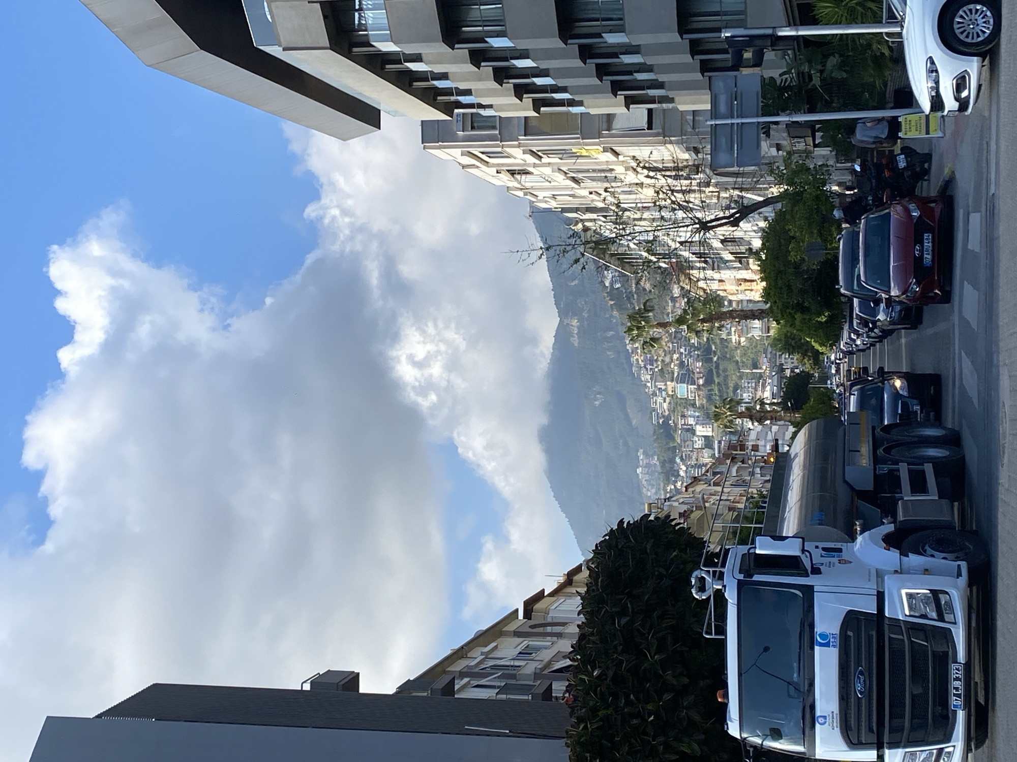 An urban street scene unfolds under a partly cloudy sky, featuring a bustling road lined with buildings, parked vehicles, and a panoramic view of mountains and a distant town in the background, consistent with a location like Alanya, Turkey. The image is captured during the daytime, with clear visibility and bright light.
In the foreground, a white Ford tanker truck is prominently parked on the right side of the street. Its license plate reads "07 CJB 323." The truck bears the "ASAT" logo and what appears to be "Antalya Büyükşehir Belediyesi" (Antalya Metropolitan Municipality) text, indicating its municipal purpose. Further down the street, several cars are parked, including a red car with the license plate "07 ARU 484." A person is seen sitting on a scooter or motorcycle, and another individual is visible walking in the distance. A yellow sign partially reads "WESTERN..." and "MONEY TRANSFER," suggesting a financial service.
The street is flanked by buildings of varying architectural styles. On the left, a modern building with a distinctive, blocky, multi-level facade is visible. Further along the street and on the opposite side, other multi-story residential or commercial buildings with lighter-colored facades line the road. Some trees and bushes are interspersed along the sidewalk and between buildings.
In the midground and background, majestic mountains rise, partially shrouded by large white and grey clouds. A dense collection of buildings, forming a city or town, can be seen sprawling across the mountain slopes and valleys, indicating an extensive settlement in a hilly coastal region. The sky above is a mix of bright blue patches and significant cloud cover.