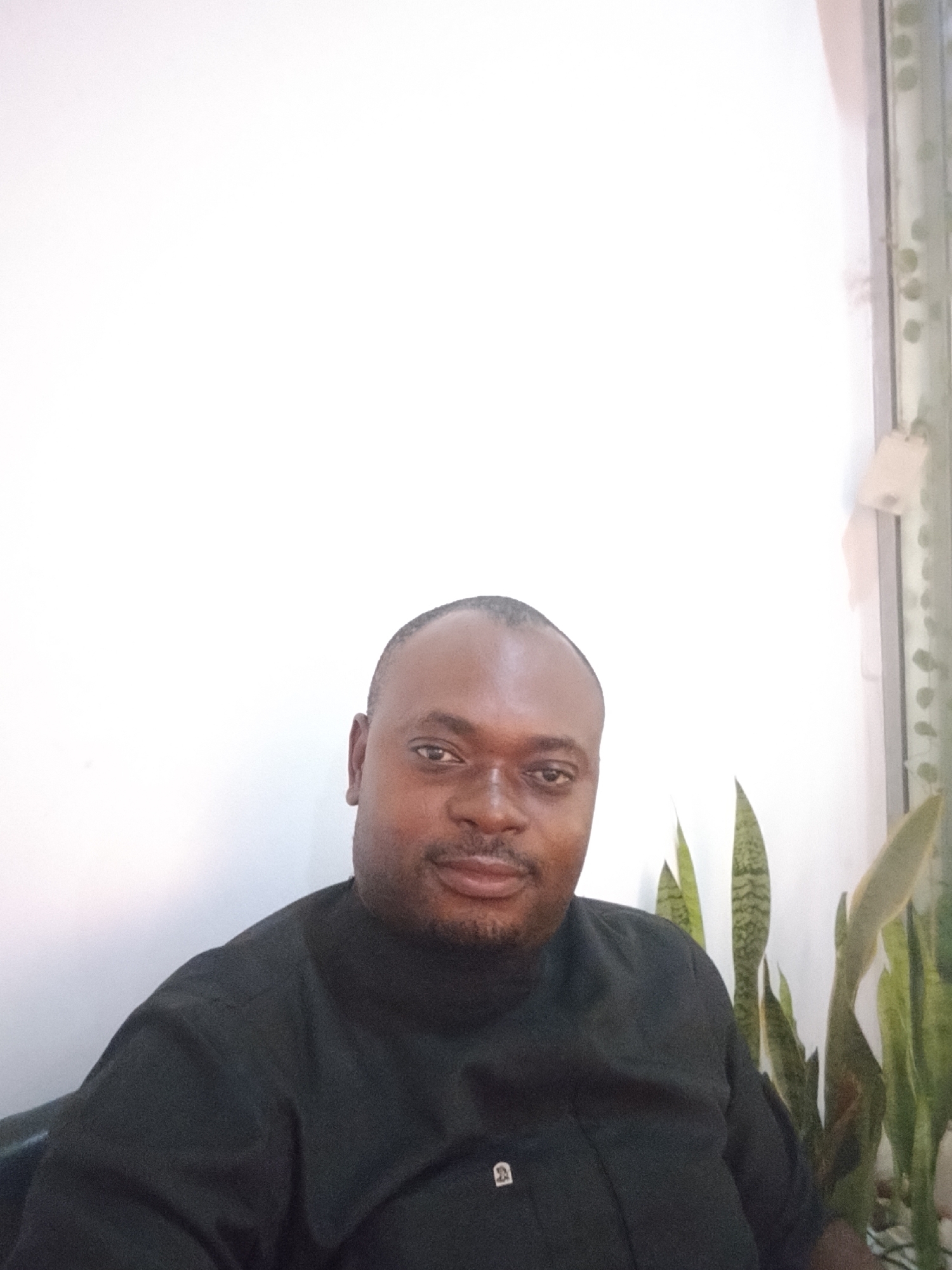 A man is seated indoors. He has dark skin, a shaved head, and a goatee, and is wearing a black shirt with a small square pin or emblem on the chest. Behind him is a plain white wall, and to his right is a cluster of tall, striped snake plants. The location is inferred to be Gwarinpa, Nigeria, based on the prompt.
