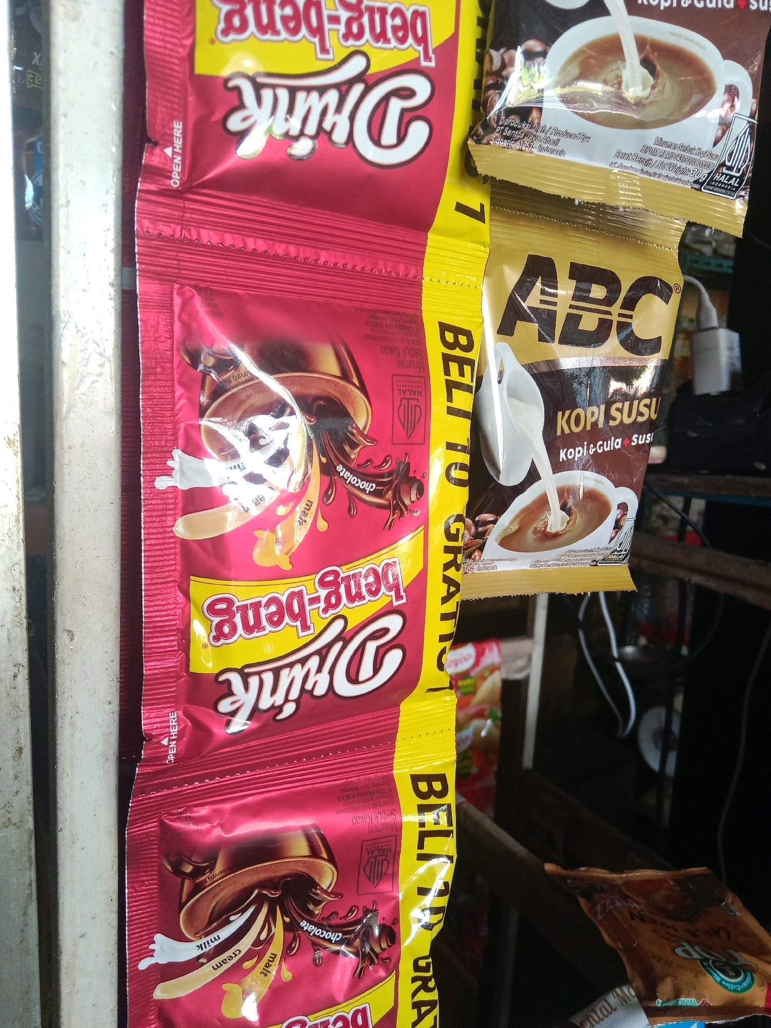 The image shows a close-up of several packets of instant coffee and chocolate drink mix hanging vertically. The packets are colorful, with dominant red, yellow, and gold colors.
The left-most packet, which is red, features the branding "Drink Beng-beng" in white and pink lettering, with illustrations of chocolate, milk, and cream. This packet appears to be a chocolate drink mix. An "OPEN HERE" tab is visible on the left side of this packet.
To the right of the red packet is a yellow packet with "BELI 1 GRATIS" (Buy 1 Get 1 Free) written vertically in black. This packet is partially obscured by the red packet.
The next packet, which is gold, displays the "ABC" logo prominently in black letters. Below the logo, it reads "KOPI SUSU" (Coffee Milk) and "Kopi + Gula + Susu" (Coffee + Sugar + Milk), indicating it is an instant coffee and milk mix. There is an illustration of milk being poured into a cup of coffee. A "HALAL" logo is visible on this packet.
Further to the right, another gold packet with the "ABC" logo is visible, also likely an instant coffee mix. There are also glimpses of other packets and products in the background, including what appears to be another product with "LA" and "ZARA" visible.
The packets are likely displayed for sale in a small shop or market stall. The context suggests this scene is located in Kertek, Indonesia, where such products are commonly sold. The lighting is somewhat dim, suggesting an indoor or shaded environment. The overall impression is of a retail display of convenience food and beverage products.