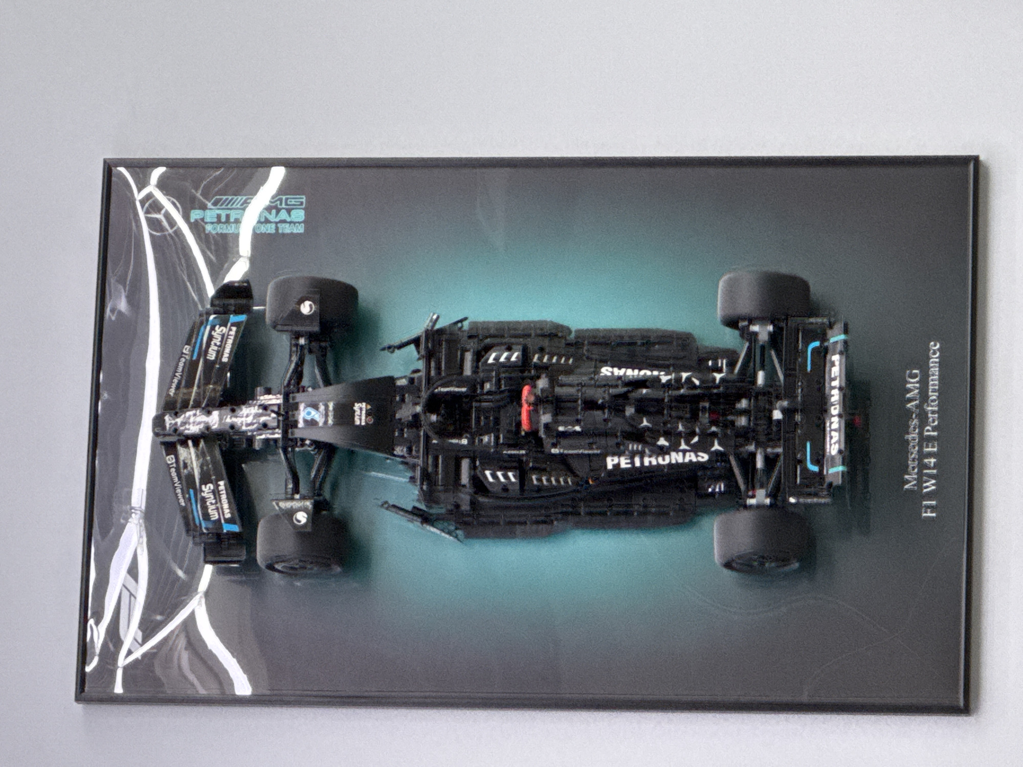 A framed, top-down view of a black and silver Formula 1 race car model is presented against a dark gray and teal background. The car is highly detailed, featuring numerous sponsor logos, including "PETRONAS," "TeamViewer," and "SYNCIUM." The "Mercedes-AMG F1 W14 E Performance" text is visible at the bottom of the frame. The framing itself appears to be a dark, possibly black, material. The photograph is taken from an elevated, slightly angled perspective, showcasing the car's intricate design. There is no indication of location in Saint Petersburg, Russia.
