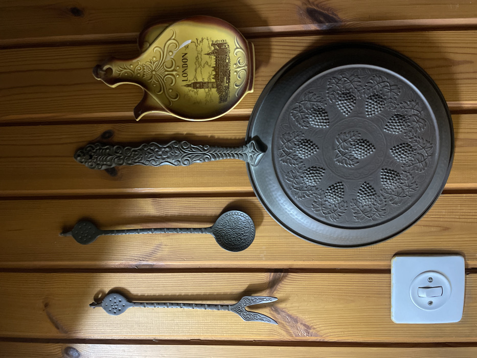 The scene depicts a section of an interior wall in Petah Tikva, Israel, constructed from vertically oriented light-toned wooden planks displaying distinct grain and knots. Four decorative objects and one electrical switch are mounted on the wall.
From left to right, top to bottom:
1. A dark metallic, ornate two-tine fork-like utensil hangs from a hook. Its handle features a textured, circular top section.
2. Adjacent to the right, a dark metallic, ornate spoon-like utensil hangs from a hook. Its handle also has a textured, circular top section, and the spoon's bowl is intricately patterned.
3. To the right of the spoon, a large, round, dark metallic pan or plate is mounted. Its surface is embossed with a central cluster of grapes surrounded by eight additional grape clusters arranged radially. The pan has a decorative, scrolled handle.
4. To the far right, a yellow and brown ceramic decorative wall ornament, shaped like a stylized pitcher, hangs from a hook. It features a painted depiction of the London skyline, including Big Ben, with the word "LONDON" prominently displayed.
5. In the lower-left quadrant of the image, a white rectangular electrical wall switch plate is mounted. It contains a single toggle switch positioned in the downward or "off" state.
All hanging items are secured to the wooden wall by visible hooks. No individuals are present in the image, and no active interactions are occurring within the static scene.