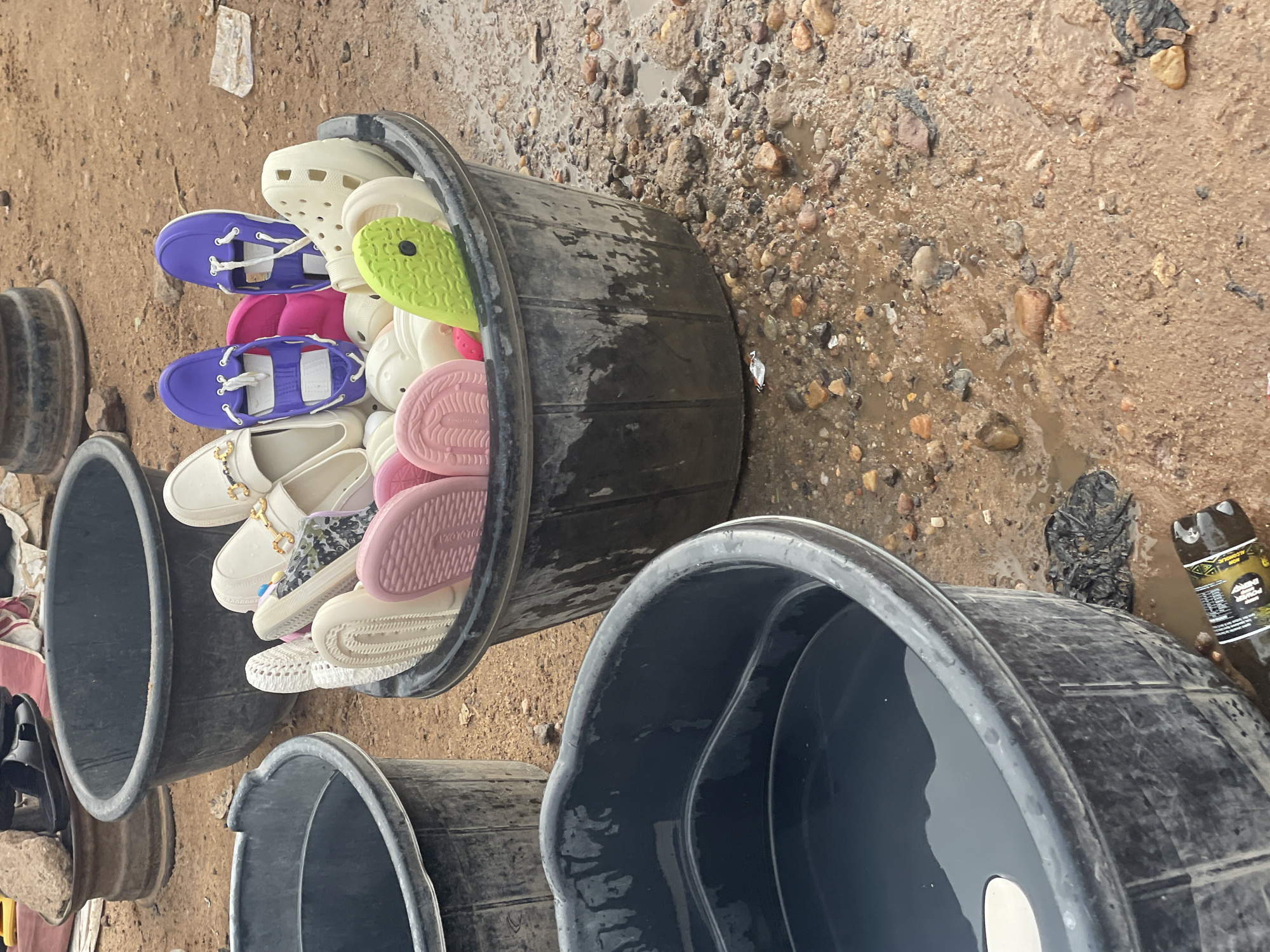 The image depicts an outdoor scene in Lokoja, Nigeria, likely a market or a roadside stall. In the foreground, a large, dark, circular plastic tub filled with water is partially visible. To its right, a black plastic tub is overflowing with a variety of colorful footwear, including white loafers with gold embellishments, purple sandals with white lacing, pink slides, and green clogs. The ground surrounding the tubs is muddy and covered with small rocks and puddles, suggesting recent rainfall. In the background, another large black tub and a metal drum are visible. The overall impression is one of an informal commercial setting, possibly selling footwear.