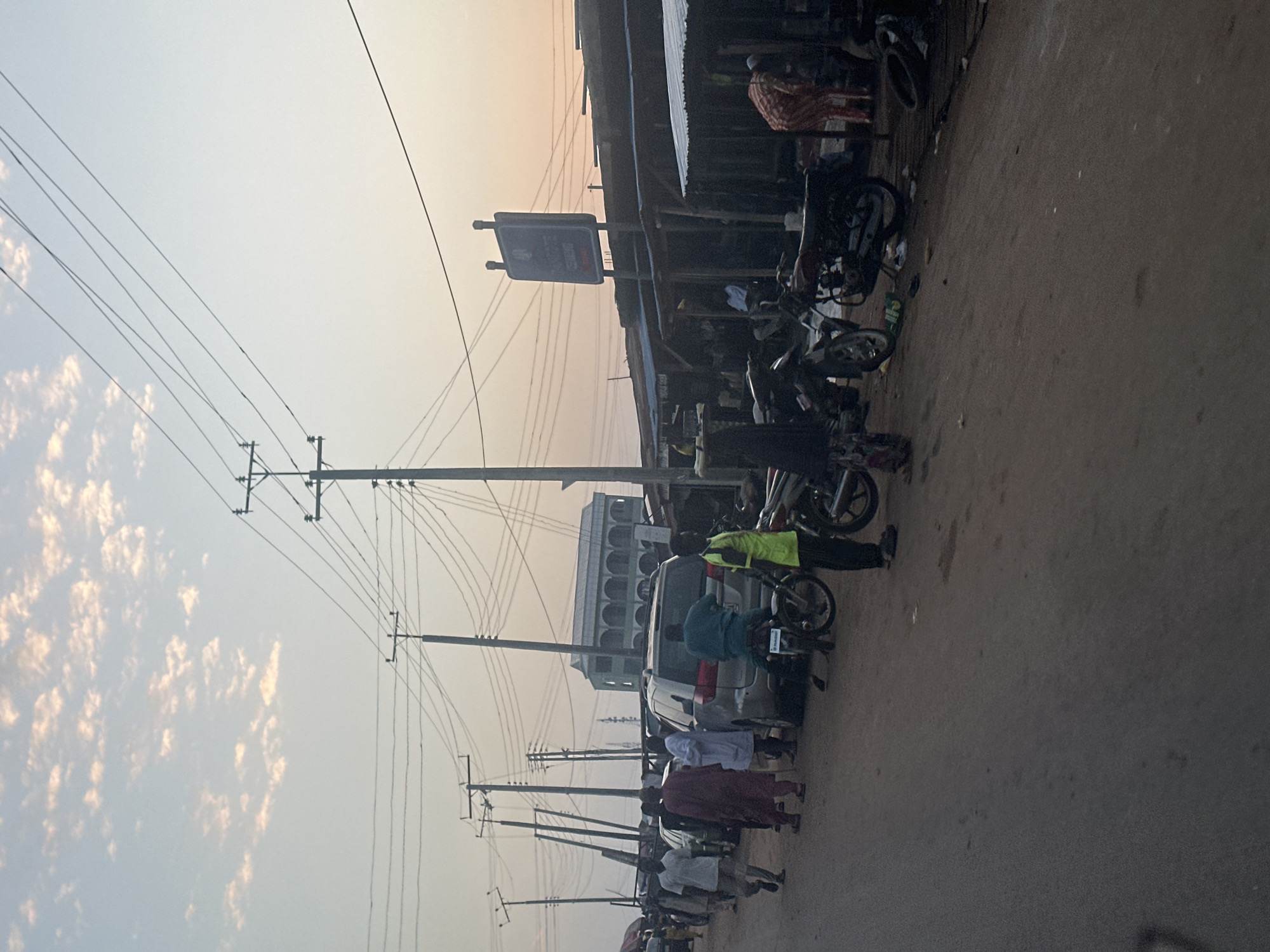 The image depicts a street scene in Akwanga, Nigeria, during what appears to be either dawn or dusk, given the soft, diffused light and pale sky with scattered clouds. The environment is dusty and unpaved, with a road surface that is dark and somewhat uneven.
Along the right side of the road, a row of makeshift market stalls or shops are visible. These structures have corrugated metal roofs and wooden supports, with some appearing weathered and rudimentary. Behind these stalls, a large, light-colored building with arched windows and a prominent roofline, resembling a mosque or a public institution, is partially visible in the background.
A significant number of utility poles with a complex network of overhead wires dominate the upper portion of the frame, indicating an established infrastructure for electricity or telecommunications.
The street is populated by several individuals and a mix of vehicles. People are dressed in casual attire, with some wearing traditional garments. Several motorcycles, including some parked and others potentially in use, are present. A silver minivan is parked in the middle of the street, and other cars are visible further down the road.
The overall atmosphere suggests a busy, functional area of the city, likely a commercial or transportation hub. The scene is captured at a low angle, giving a sense of immersion in the environment.