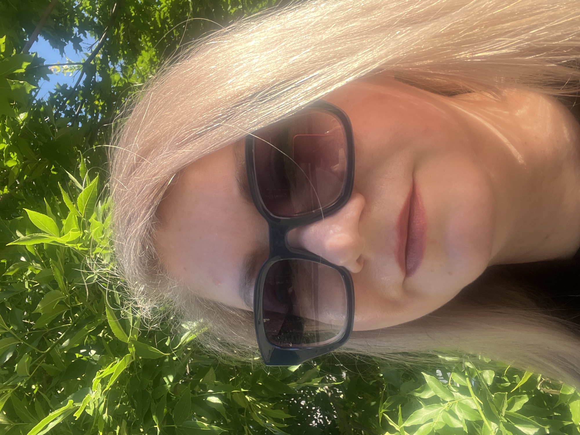 A close-up, head-and-shoulders selfie of a fair-skinned woman with blonde hair wearing dark sunglasses and subtle lipstick. She is positioned in front of a background of lush green foliage and a clear blue sky, suggesting an outdoor setting. The lighting indicates it is daytime, with sunlight illuminating her face and hair. The image does not provide any identifiable landmarks or clues to determine its location in Izhevsk, Russia.