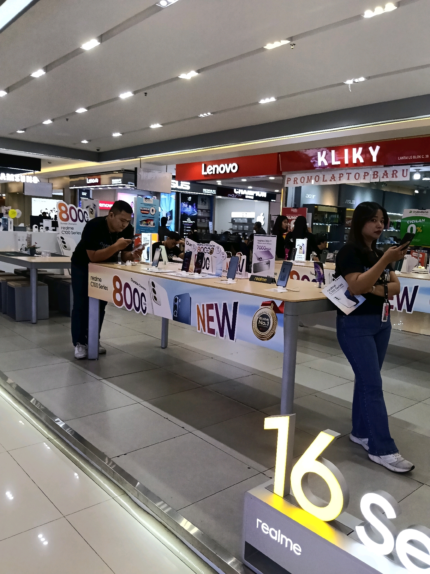 In a retail electronics store in Tangerang, Indonesia, a man in a black t-shirt and dark pants stands at a display table, looking at his phone. He is wearing white sneakers. He leans on the table, which features promotional material for "realme C100 Series" and "8000mAh." Several smartphones are displayed on stands on the table. A woman in a black t-shirt and blue jeans stands to the right, also looking at her phone. She is wearing white sneakers. Behind the tables, various electronic store displays are visible, including signs for "Lenovo" and "KLIKY PROMO LAPTOP BARU." Large illuminated numbers, "16," and the word "realme" are visible on the floor in the foreground. The store environment appears well-lit with fluorescent lighting on the ceiling.