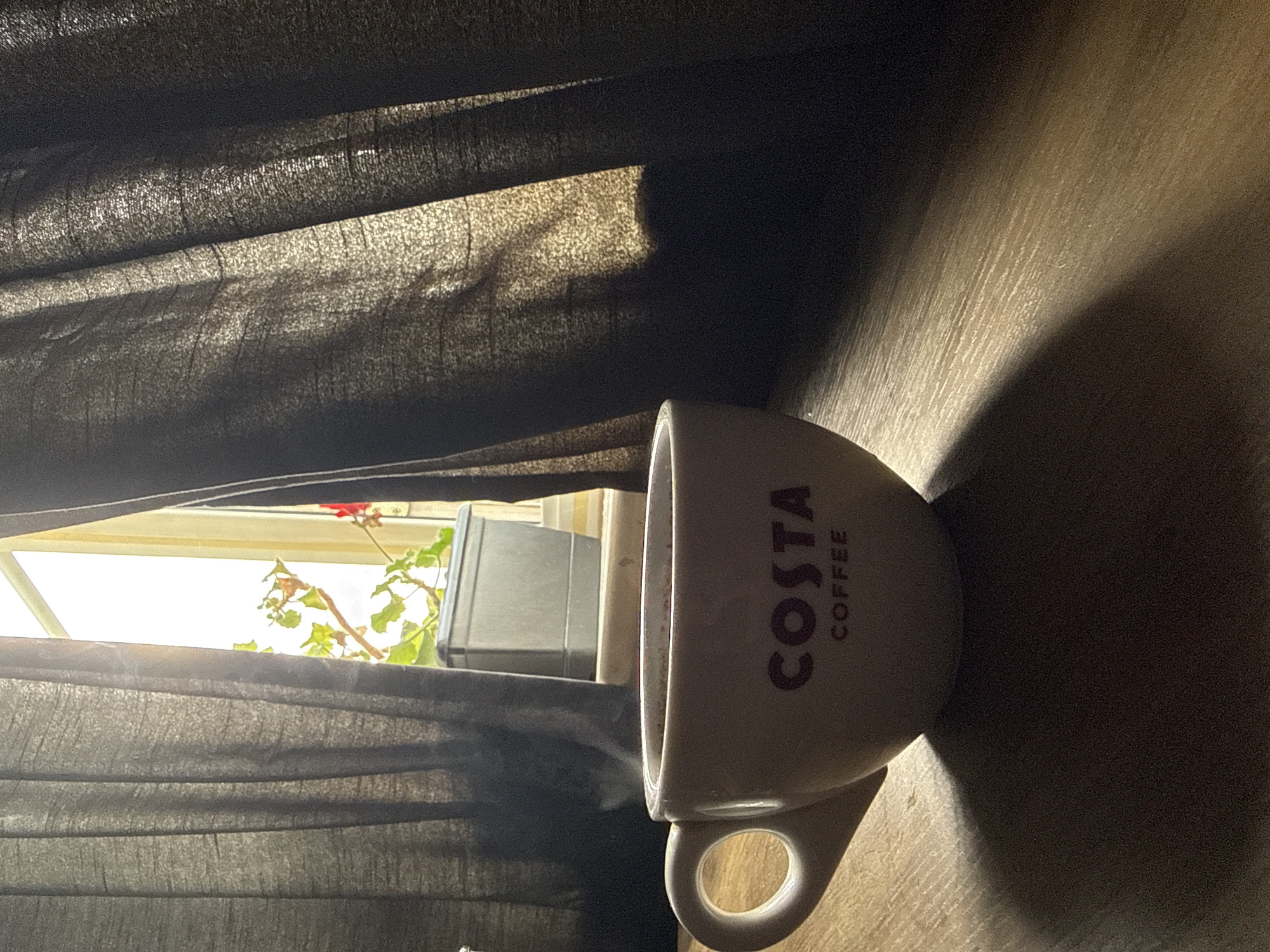 The image displays an indoor scene during daylight hours in Great Yarmouth, United Kingdom. A white ceramic mug with "COSTA COFFEE" printed in dark maroon text sits on a light-colored, textured wooden or laminate surface, positioned on the right side of the frame. The mug casts a distinct, elongated shadow across the table surface.
To the left, dark grey, textured curtains are drawn, creating prominent folds. Strong natural sunlight filters through and around the curtains, illuminating their fabric and the table. Through a gap in the curtains, a brightly lit window is visible, revealing part of a vibrant green plant with small red flowers, likely a geranium. A grey, rectangular object, possibly a planter or container, is also seen outside the window. The bright outdoor light suggests a sunny day, creating high contrast between the illuminated areas and the deep shadows cast by the curtains and the mug. The scene conveys a quiet, contemplative atmosphere with a focus on light and shadow.