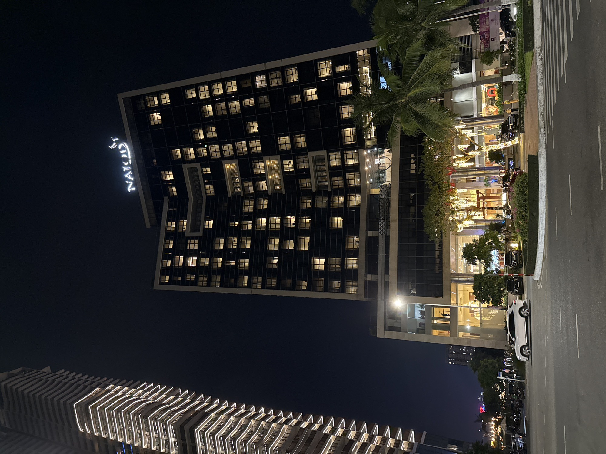 This is a nighttime photograph depicting a modern urban landscape in Đà Nẵng, Vietnam. The scene is dominated by brightly lit buildings, with a dark sky overhead.
In the upper central and right portion of the image, a tall, dark-colored skyscraper stands prominently. Its facade is characterized by a grid of numerous square windows, many of which are illuminated from within, suggesting activity or occupancy. A large, glowing white sign is partially visible on the side of this building, displaying the text "NATUOY".
Below and to the right of this skyscraper, a vibrant commercial or hotel entrance area unfolds. This zone is heavily decorated with an array of bright, warm-toned lights, possibly string lights or festive installations, and features several tall palm trees. Vehicles, including a white SUV and multiple motorcycles, are parked or moving along the adjacent road, which also shows a distinct pedestrian crosswalk. Faint figures of people can be seen moving within this brightly lit area.
In the lower-left section of the photograph, another contemporary building showcases a distinctive architectural design. This structure features a series of horizontal, wave-like elements, each illuminated with bright white strip lighting, creating a striking visual pattern against the night sky. The overall impression is one of a bustling, well-lit city center at night.