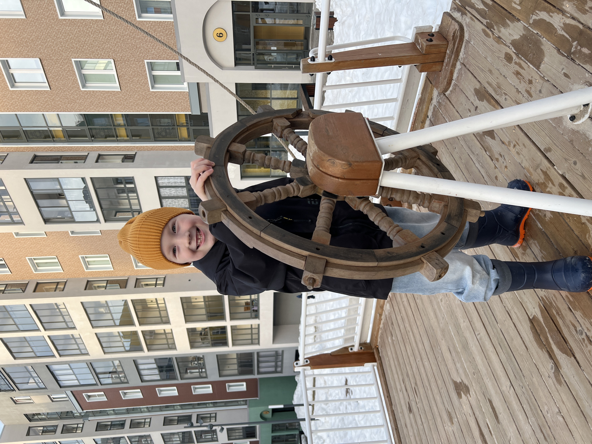 A young child, approximately 5-7 years old, is standing on a wooden deck, holding a large, dark brown wooden ship's wheel. The child is wearing a yellow knit beanie, a black jacket, light blue jeans, and dark blue boots with orange soles, and is smiling towards the camera. The wooden deck surface shows visible grain and some scattered moisture. White railings with wooden posts delineate the edge of the deck. In the background, several multi-story modern residential buildings are visible, constructed with light beige, brick-colored, and grey facades featuring numerous windows. Snow covers the ground surrounding the deck. A specific building entrance, marked with a yellow circle containing the number "6", is present on the right, featuring an arched entryway and glass doors, with a thick rope descending near it. The scene is located in Pervomaysky, Russia.