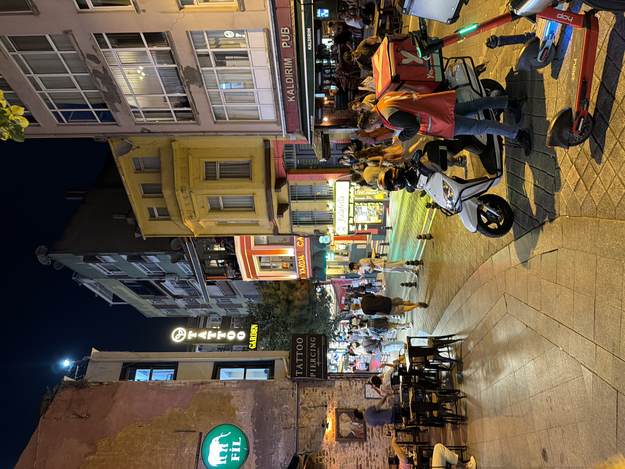 This image captures a bustling street scene at night in Kadıköy, Turkey. The street is lined with buildings featuring vibrant facades and illuminated windows. On the left, a building with weathered pink walls has a green circular sign with a white elephant and the text "FIL" below it. Adjacent is a vertical sign for "TATTOO GARDEN" and another for "TATTOO PIERCING accessories." Outdoor seating with tables and chairs is set up on the sidewalk in front of these establishments.
Further down the street, a yellow building stands out with intricate architectural details. To its right, a building with a red awning displays the sign "KALDIRIM PUB." Several people are visible on the street, walking, sitting at tables, and standing. A white scooter with a red delivery box is parked in the foreground, and a red electric scooter is positioned next to it. The street is paved with patterned tiles, and streetlights cast long shadows. The overall atmosphere suggests a lively urban environment in the evening.