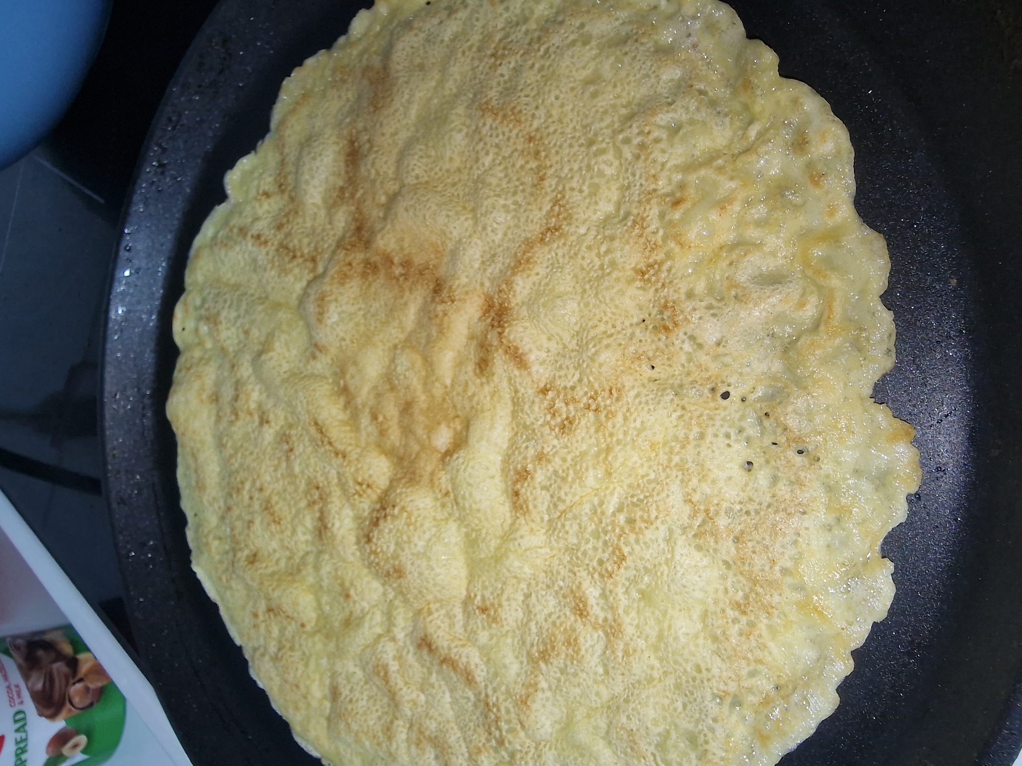 The image shows a large pancake being cooked in a black frying pan. The pancake has a bubbly surface with some browning. The pan appears to be on a stovetop, with a blue bowl visible in the background and a package of hazelnut spread partially visible in the lower left corner. The setting is a kitchen, but the specific location is unknown.