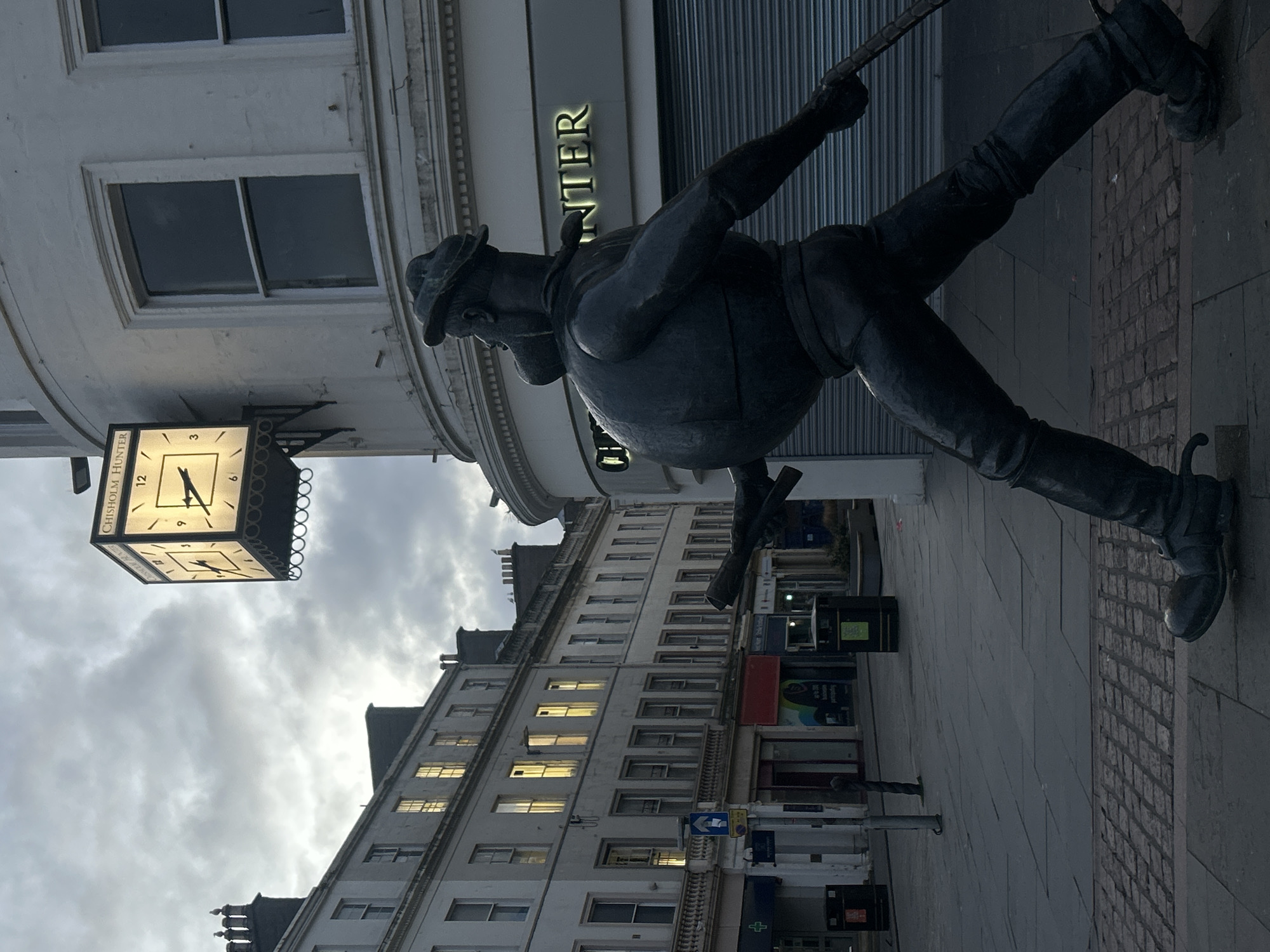 An urban street scene in Dundee, United Kingdom, captured from a low, upward-angled perspective, revealing buildings, a prominent statue, and an overcast sky.
The central subject is a dark, bronze-colored statue of a stout man, depicted in mid-stride with his left leg raised. He wears a flat cap, a jacket or waistcoat, and sturdy boots. He holds a long, slender object, possibly a whip or stick, in his right hand which extends upwards. The statue appears to be positioned on a paved sidewalk, part of which shows a brick pattern.
To the left of the statue, a square, ornate illuminated clock hangs from a building facade. The clock face shows the time as approximately 5:30. Partially visible text on the clock reads "CHISHOLM H". Behind the statue, large, illuminated gold letters spelling "NTER" are seen on a building, which, combined with the clock's inscription, strongly indicates "Chisholm Hunter", a jeweller.
Further in the background, a multi-story building extends along the street, featuring numerous windows. Some of these windows show warm yellow light from inside, suggesting either evening or an overcast day. The sky above is uniformly grey and cloudy. The overall setting is a bustling street, possibly a shopping or pedestrian area, characterized by its architectural details and public art.