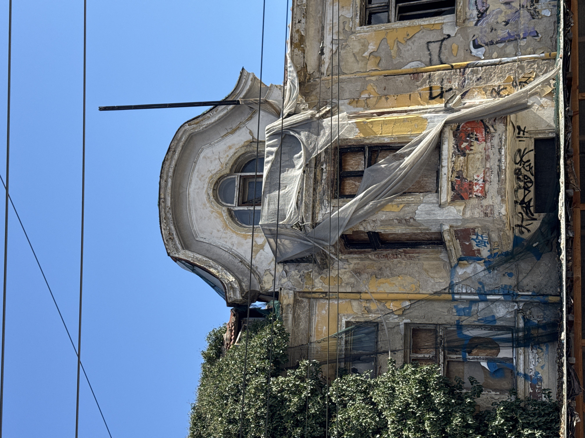 The image depicts the dilapidated facade of a building in Sofia, Bulgaria. The structure shows signs of disrepair, including peeling paint, graffiti, and crumbling plaster. The building's architectural details, such as a rounded window and ornate molding, suggest a once-grand design. A translucent sheet is draped over a portion of the building. Vegetation grows on the building at the base. Electrical wires run across the blue sky in the background.