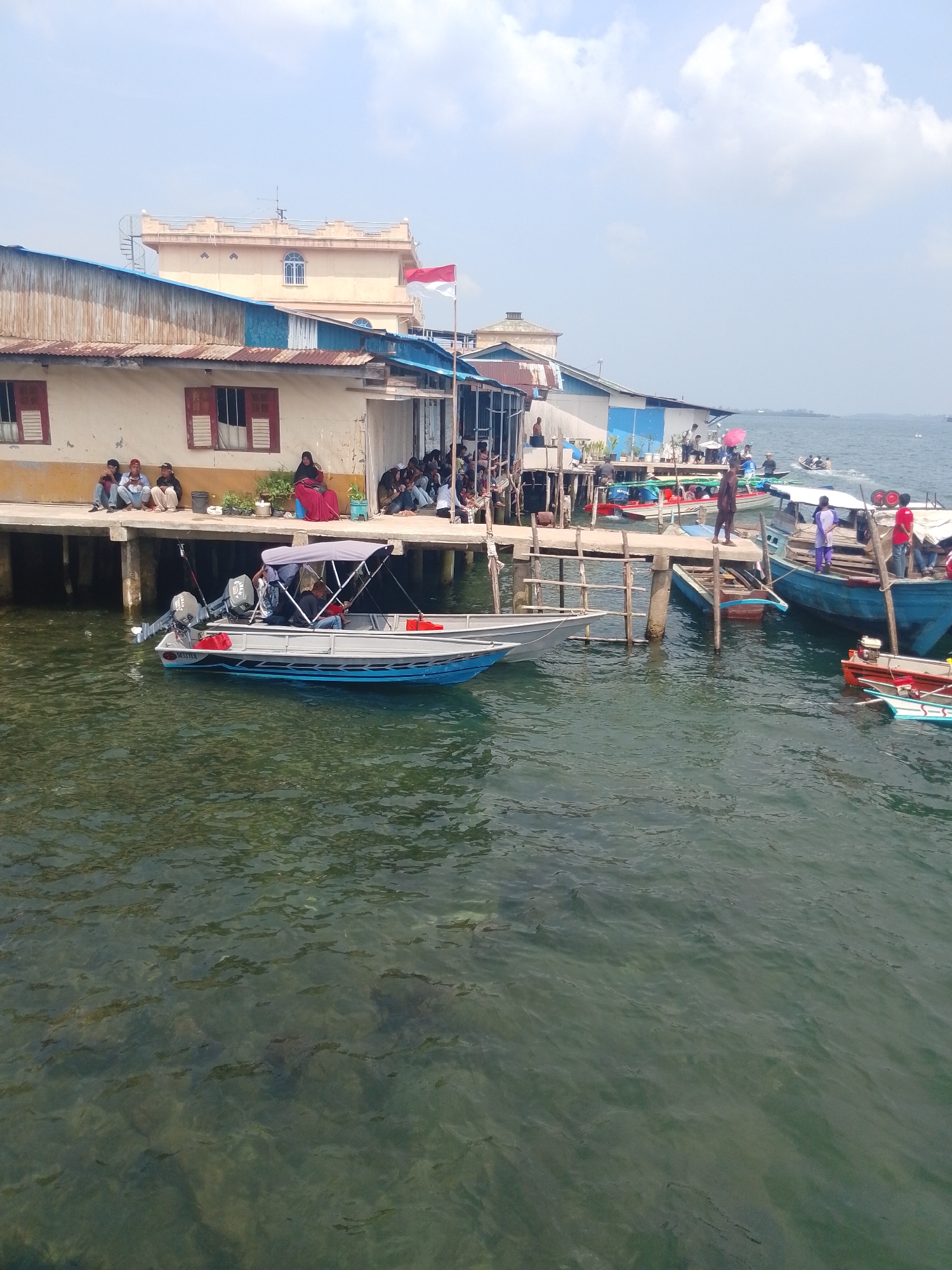 The image depicts a waterfront scene in Batam, Indonesia, likely at a dock or pier. In the foreground, a light-colored motorboat with a canopy and two outboard motors is moored in clear, greenish water. The boat has blue trim along its waterline and red accents on its sides. Several people are visible on a wooden platform extending from a building along the water's edge. Some are seated, while others are standing. The building appears to be a single-story structure with corrugated metal roofing and painted walls, featuring several windows with red shutters. Further back, more boats are docked, including a larger blue vessel with people on board and several smaller, colorful boats. In the background, a multi-story building with a light-colored facade and decorative architectural elements is visible. An Indonesian flag is flying from a flagpole in front of this building. The sky is blue with scattered white clouds. The overall impression is a bustling water-transport hub or a residential area situated along the coast.