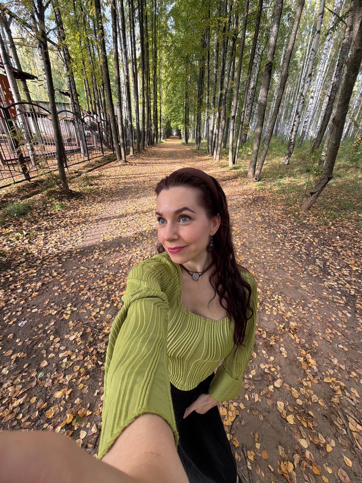 In Chaikovsky, Russia, a woman with long, dark wavy hair and blue eyes stands on a path in a park during autumn. She is wearing a green ribbed top with a square neckline and a black skirt. Her left hand is placed on her skirt. She is looking to her right with a slight smile. The path is covered with fallen yellow leaves, and tall, slender trees line both sides of the path, creating a tunnel-like effect. A metal fence is visible on the left side of the path, and beyond it are more trees and some structures. The sunlight filters through the trees, casting shadows on the ground.