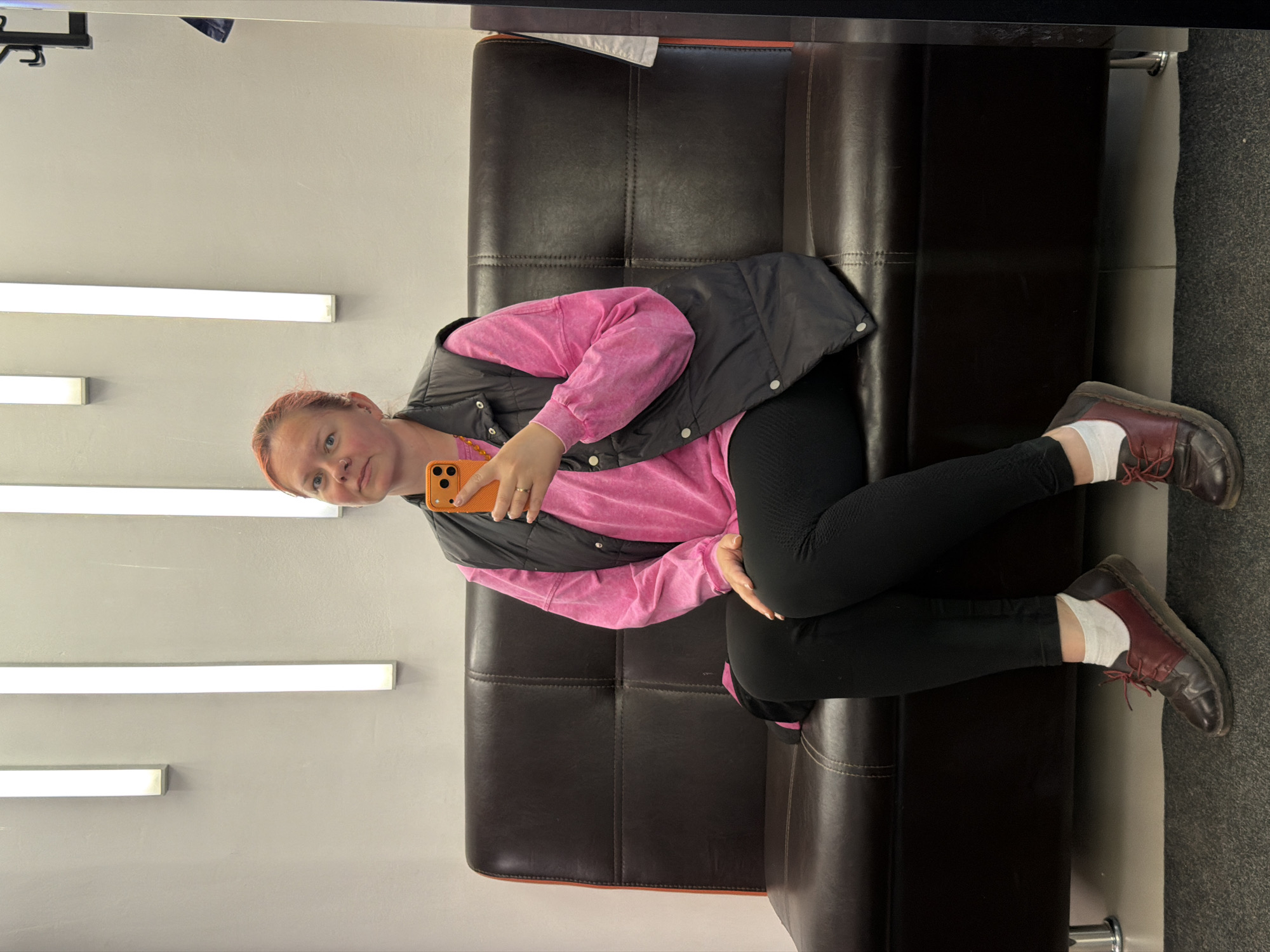 A woman is lying on a dark brown leather couch with chrome legs. She is wearing black leggings, a pink long-sleeved shirt, a black vest, white socks, and brown and red leather shoes. She is holding an orange smartphone in her right hand, taking a selfie. Behind her is a light gray wall with several long, thin, illuminated light fixtures. The floor to the right of the couch is gray. The location is likely in Chaikovsky, Russia, based on the image context.