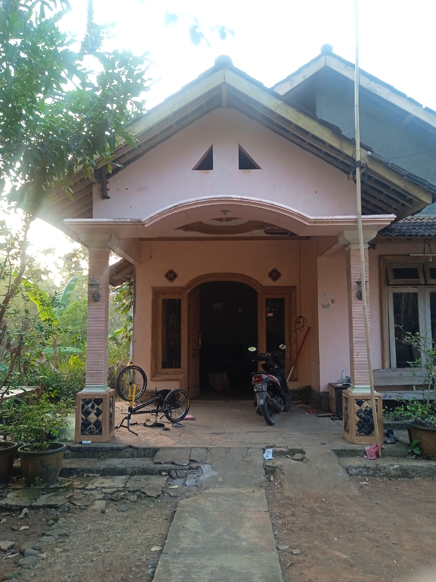 The image depicts the front of a single-story house with a light pink facade, located in Jatisrono, Indonesia. A paved pathway leads to the entrance, which is framed by an arched doorway and flanked by two large windows. The house has a sloped roof covered with dark tiles. A covered porch area is supported by fluted pillars.
In front of the house, a black motorcycle is parked. To the left of the entrance, a bicycle is propped up on a stand, with its wheels visible. Several potted plants are placed on the ground near the pathway and the porch. The surrounding environment appears to be a rural or suburban area, with lush green trees and foliage visible in the background. The sunlight is bright, casting shadows and creating lens flare effects in the upper left portion of the image. The ground is a mix of dirt, cracked pavement, and concrete. A broom or long pole leans against the house near the entrance.