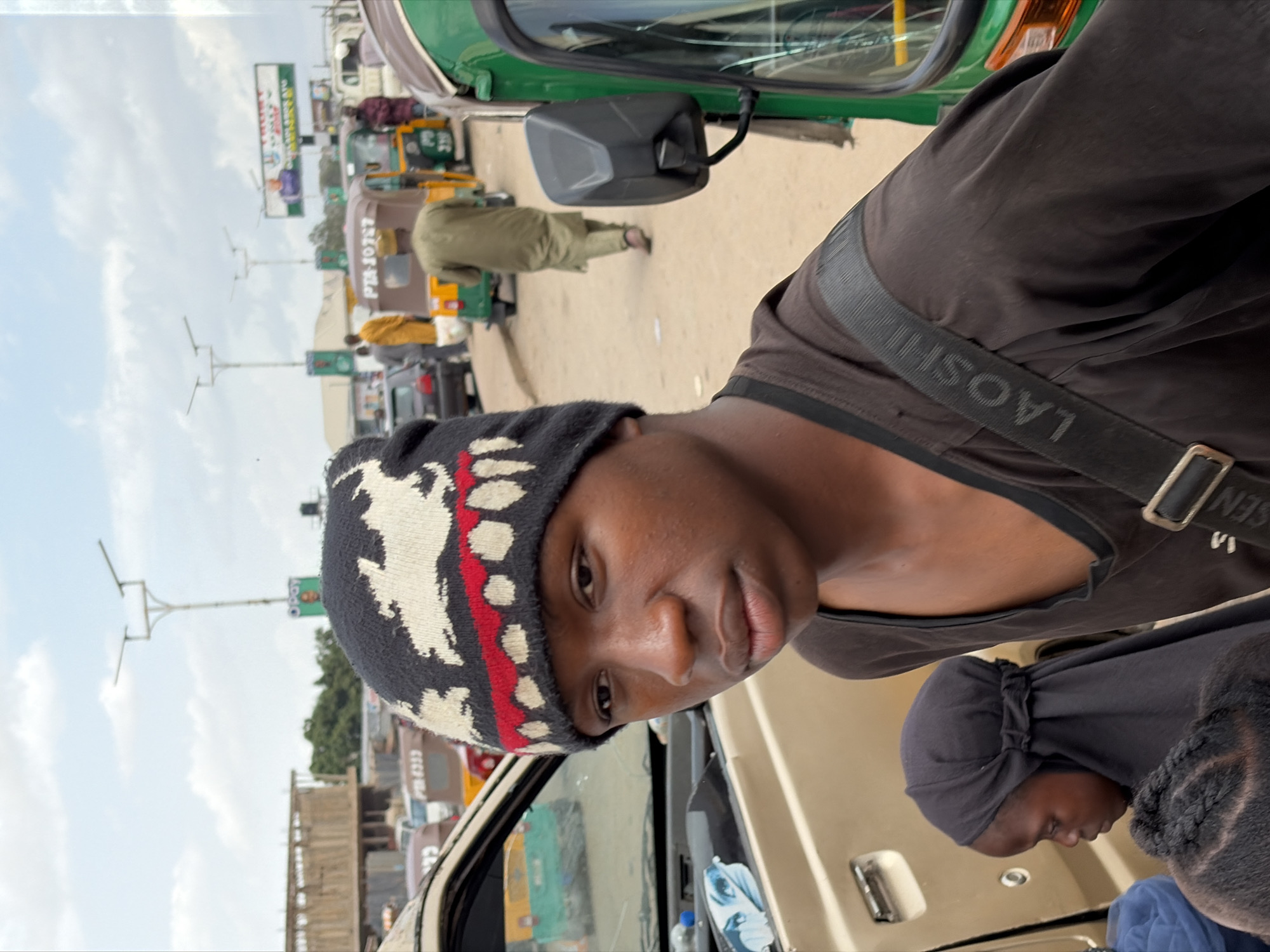 The image depicts a young man wearing a patterned beanie and a black shirt with a strap across his shoulder, seemingly taking a selfie from inside a vehicle. The background shows a bustling street scene in Gura Top, Nigeria, with auto rickshaws ("Keke Napep"), other vehicles, and people. Utility poles and billboards are visible in the distance. Another individual with a black head covering is partially visible next to the man.