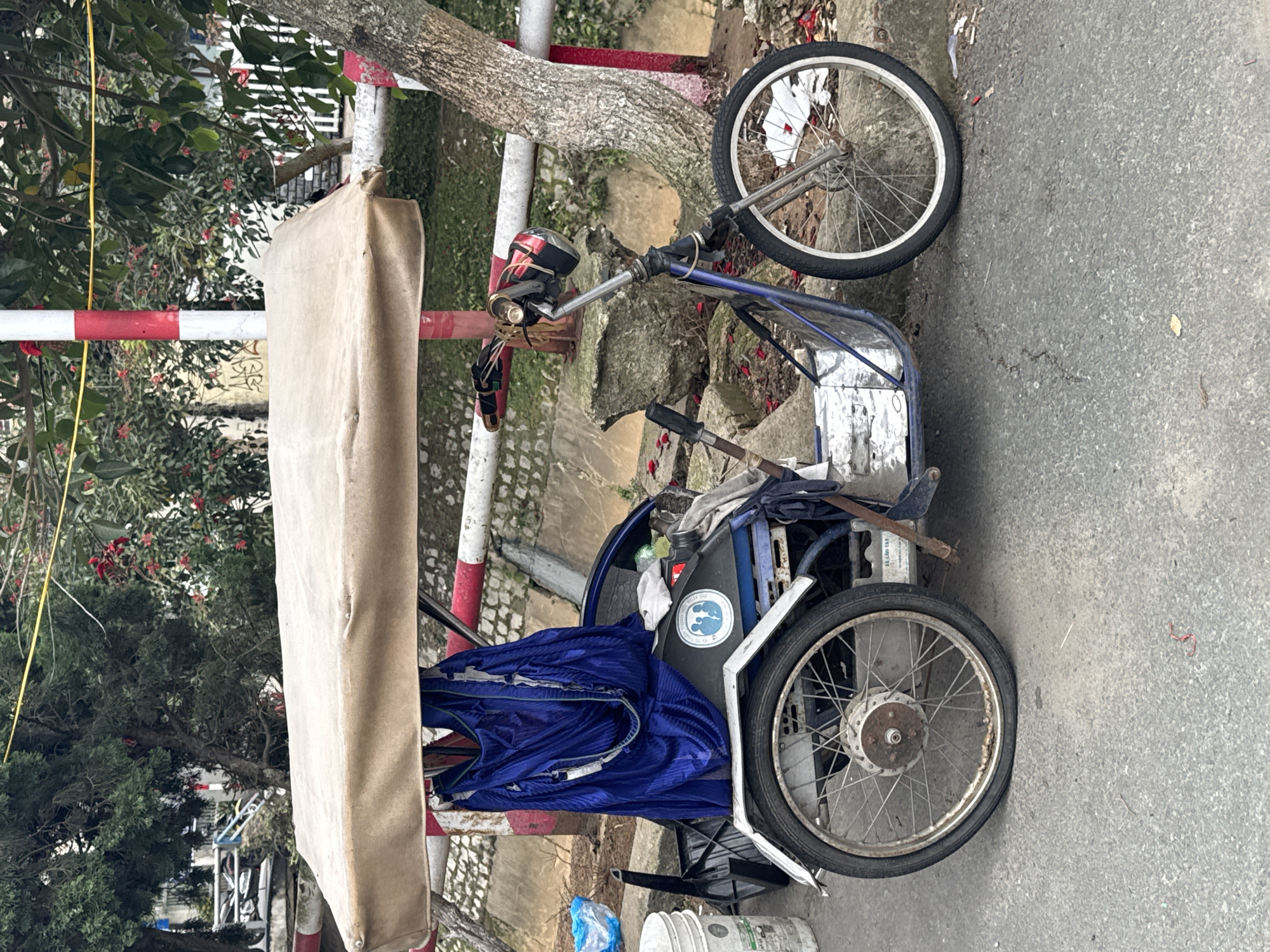 This image depicts a three-wheeled vehicle, likely a cyclo or a motor-assisted bicycle rickshaw, parked on an asphalt road in Da Lat, Vietnam. The vehicle is positioned outdoors, with a light-colored canopy extended over its seating area. A bright blue fabric, possibly a garment or covering, is draped over the frame. The vehicle has two wheels at the rear and one at the front, with a basket-like cargo area. The surrounding environment includes trees, a stone wall, and a red and white striped pole, suggestive of a roadside or park area. There are no people visible in the image. The overall impression is that of a common mode of transportation or vending vehicle in the region, temporarily parked.