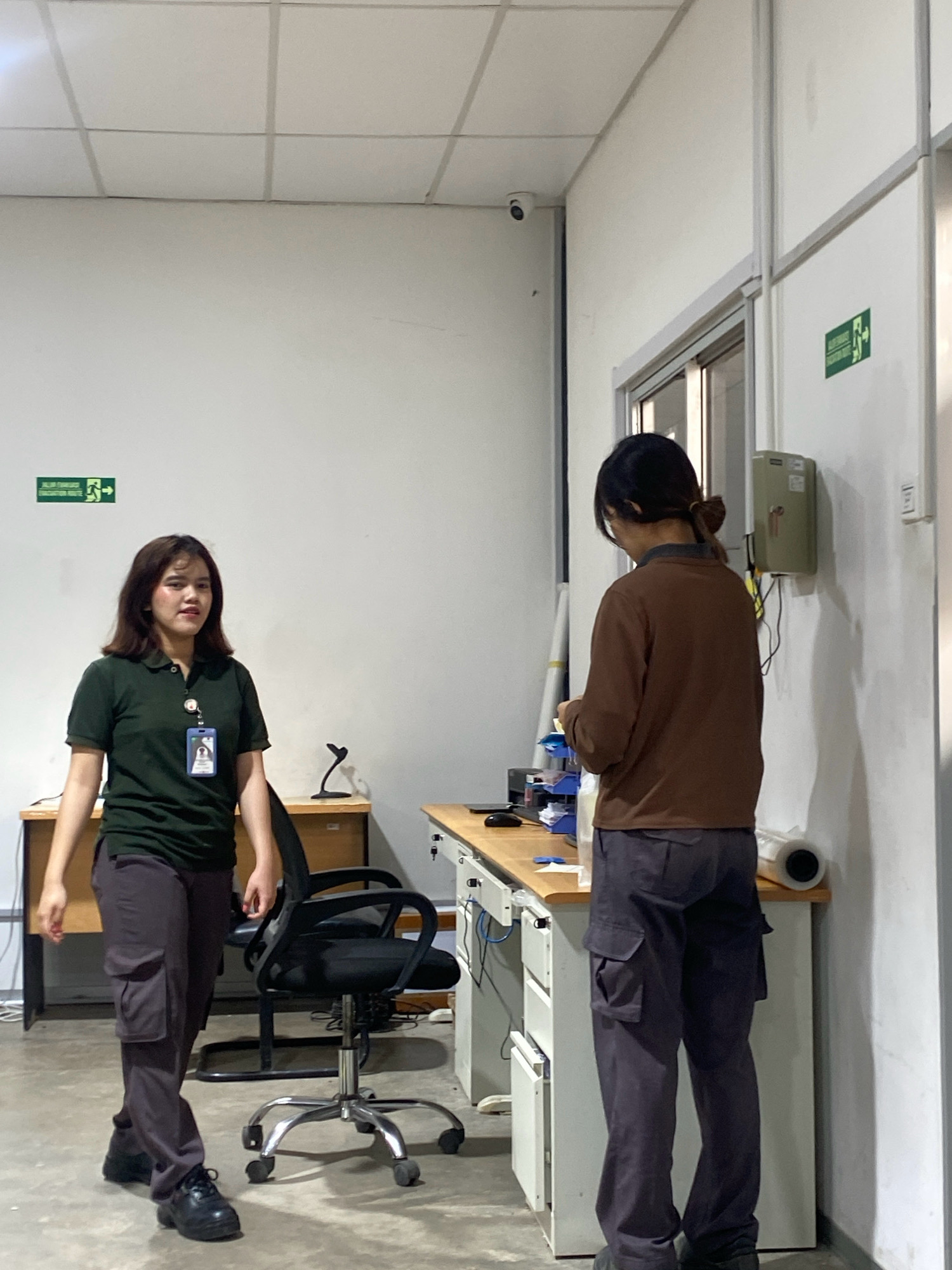 Two women are standing in what appears to be an office or workshop environment. The woman on the left is wearing a dark green polo shirt with a lanyard around her neck and dark gray cargo pants. She has dark hair and is looking towards the camera. The woman on the right is wearing a brown long-sleeved shirt and dark gray cargo pants, with her back to the camera, appearing to be examining something in her hands.
The environment includes a desk with office supplies, a rolling office chair, and a wall-mounted cabinet. There are also exit signs visible on the wall. A security camera is mounted on the ceiling. The flooring is concrete. The scene suggests an industrial or commercial setting within Batam, Indonesia.