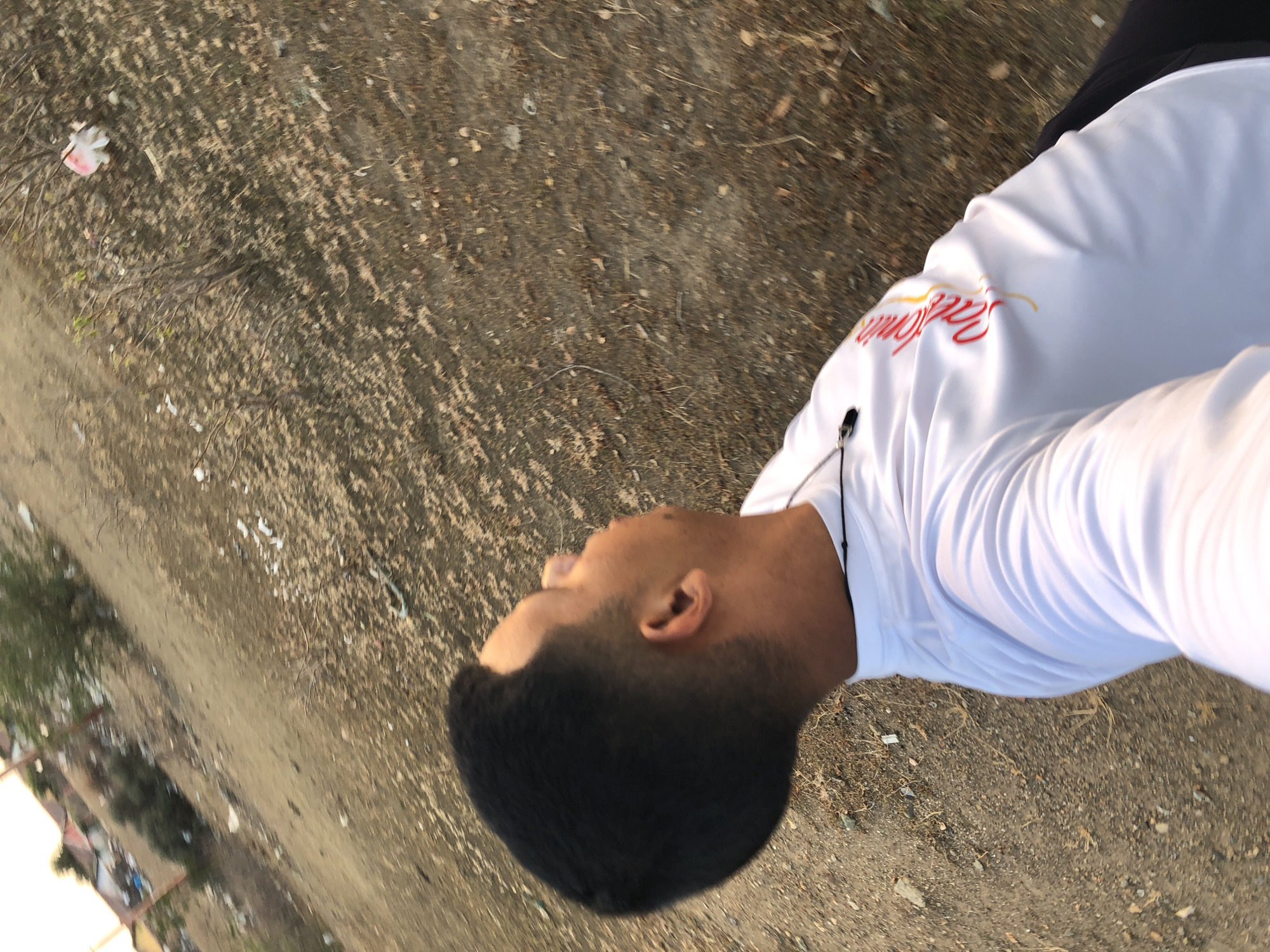 The image depicts a person with a close-cropped black haircut, wearing a white, long-sleeved shirt with red lettering on the shoulder. They are outdoors on a dirt and gravel surface. The background shows signs of debris or litter. The location cannot be definitively identified as Altagracia, Venezuela without additional information.