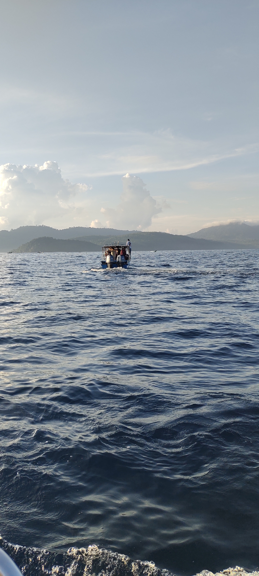 A small boat with several passengers is traveling across a body of water, leaving a wake behind it. The boat is dark blue with a lighter colored roof structure, and several people are visible in and around it. In the background, lush green hills and mountains rise from the water, with some clouds in the sky above. The water is choppy with visible waves. The scene is likely a coastal area in Sabang, Indonesia, given the tropical landscape and the type of boat commonly used for tours and transport in the region. The lighting suggests it is either early morning or late afternoon.