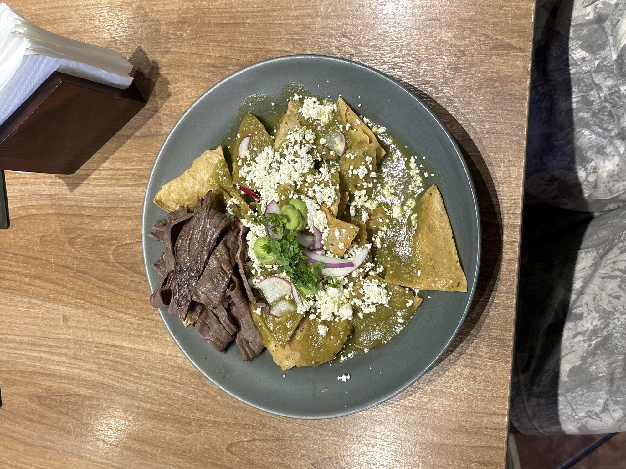 A top-down shot shows a plate of enchiladas in San Andrés Cholula, Mexico. The enchiladas, covered in green sauce and crumbled white cheese, dominate the center of the dark gray plate. Sliced radishes, red onion, and green herbs are scattered on top. A serving of sliced meat sits to the left of the enchiladas. The plate is placed on a light-brown wooden table. A napkin holder is in the upper left corner of the frame. To the right of the plate is a gray and white patterned chair.