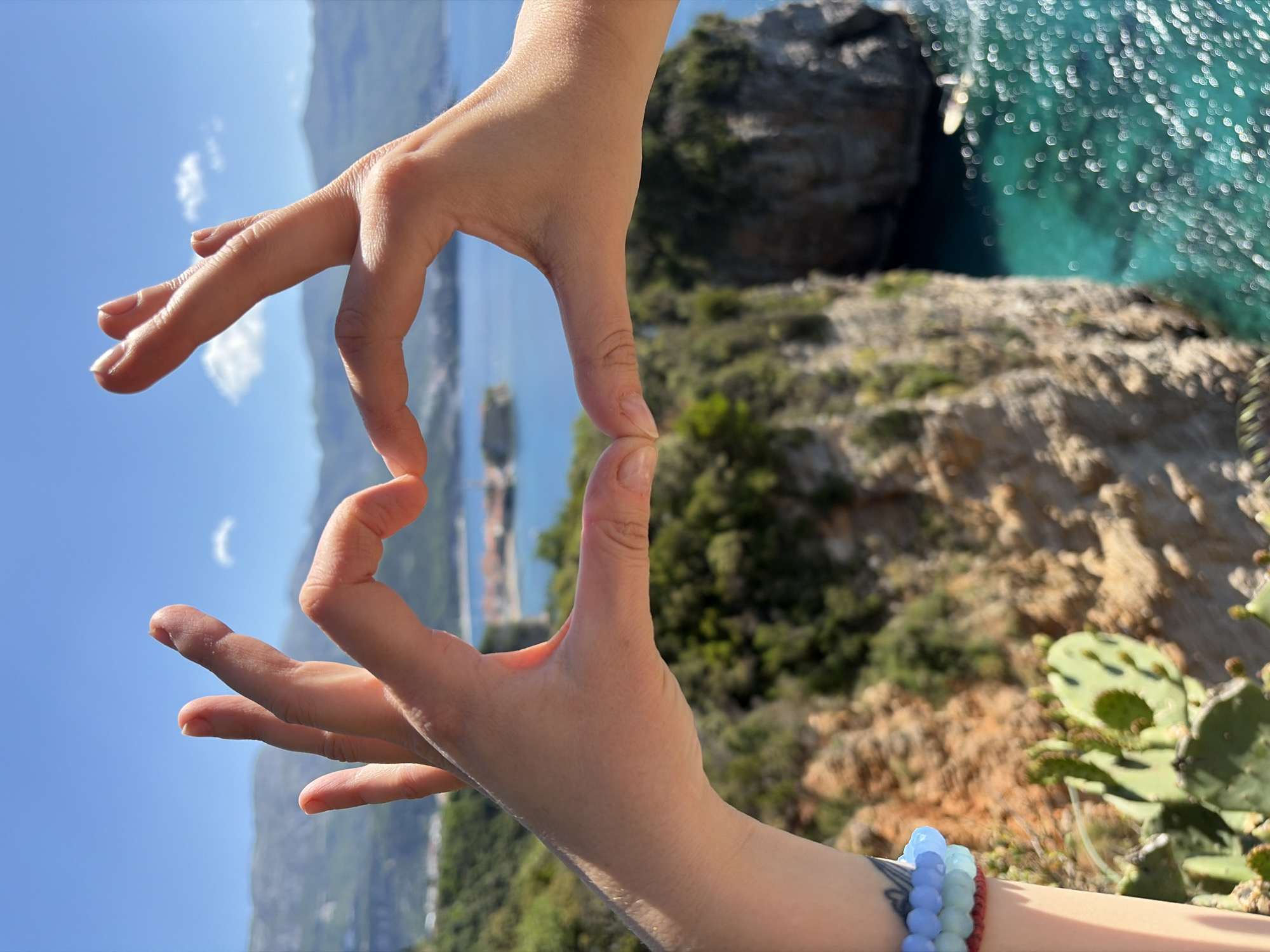 A person's hands are positioned to frame a scene. The background shows a coastal landscape, possibly near the city of Budva, Montenegro. There is water, likely the Adriatic Sea, with what appears to be a rocky island or headland in the distance. Vegetation and cliffs are visible on the coastline. The sky is blue and the weather appears to be clear and sunny. The hands are fair-skinned and one wrist has bracelets.