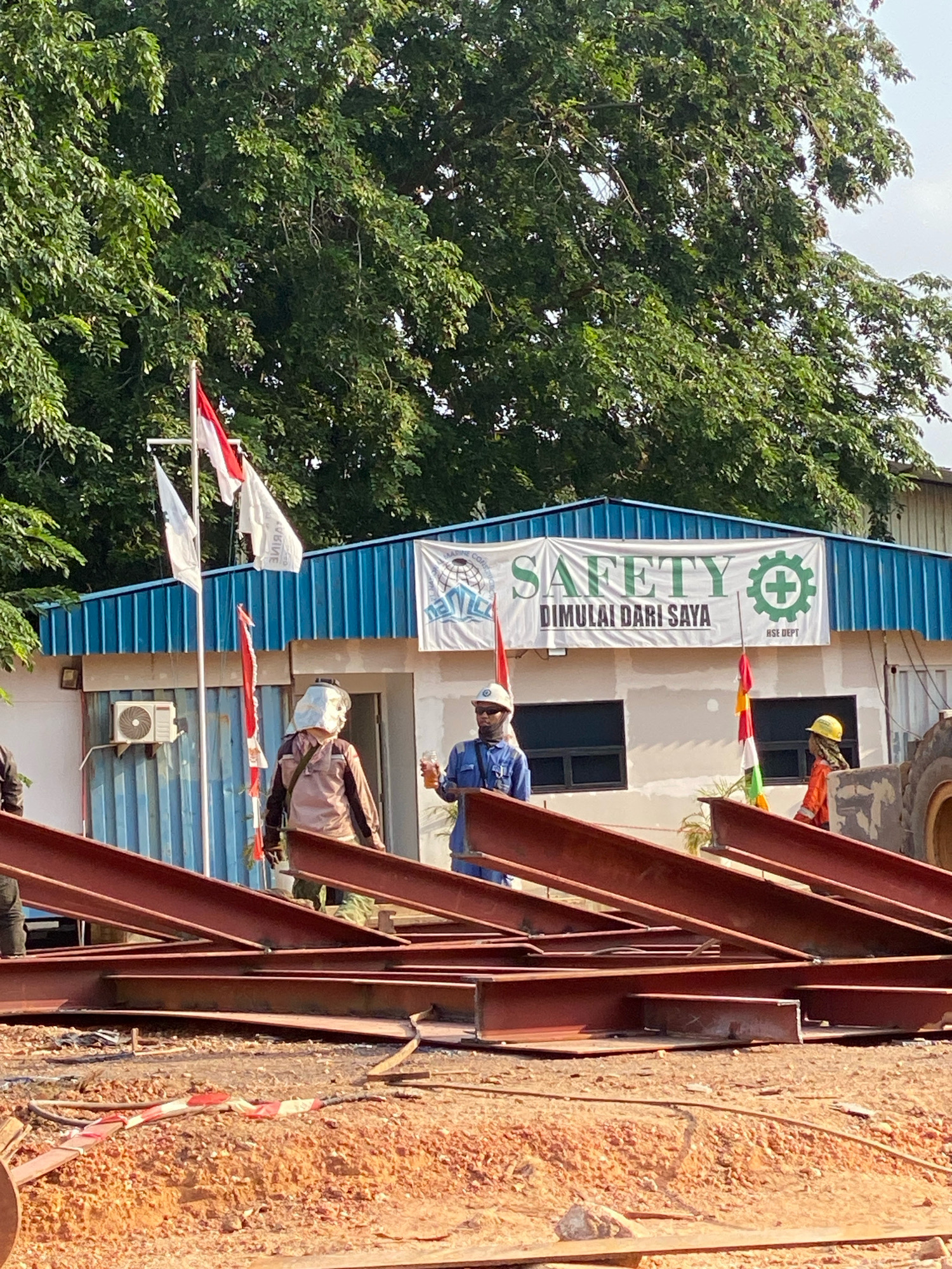 The image captures a scene on a construction site in Batam, Indonesia, with a building in the background displaying a prominent banner that reads "SAFETY DIMALAI DARI SAYA" (Safety Starts With Me) along with a green safety symbol. Two construction workers are visible; one in the foreground wearing a hard hat and safety vest, standing near a pile of large red steel beams. Another worker, dressed in a blue uniform and hard hat, is further back, holding a drink. Flags, including the Indonesian flag, are planted near the building. The environment appears to be an outdoor industrial or construction area, with trees and blue corrugated metal siding on the building. The ground is covered with dirt and construction debris, and more steel beams are stacked in the foreground.