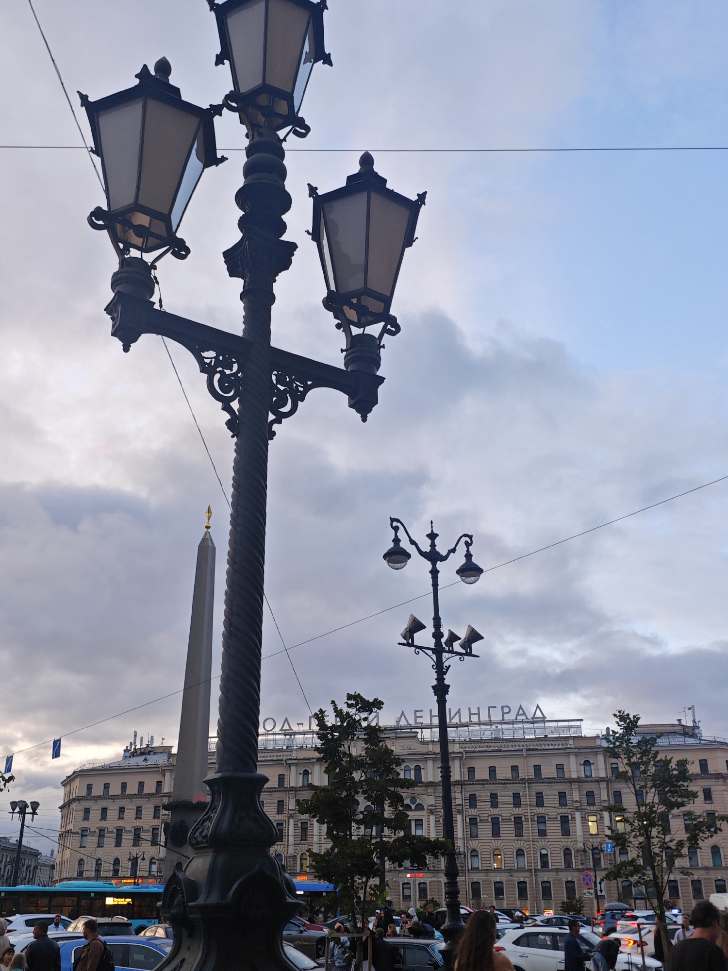In Saint Petersburg, Russia, a street scene unfolds under a cloudy sky. Dominating the foreground is a tall, ornate lamppost with three lamps at the top, its dark, twisted metal pole extending upwards. To the left of the lamppost, an obelisk with a golden spire pierces the sky. In the background, a large, light-colored building with numerous windows spans the width of the frame. Large letters spelling "ЛЕНИНГРАД" (Leningrad) are visible on the rooftop of this building. The street in front of the building is filled with cars, suggesting a busy urban environment. A blue bus is also visible on the left side of the street. Several trees with dark green foliage are interspersed throughout the scene. People are present, walking on the sidewalk and near the vehicles, but their specific actions are not discernible in detail. The overall atmosphere is that of a bustling city street in the late afternoon or early evening, with overcast weather.