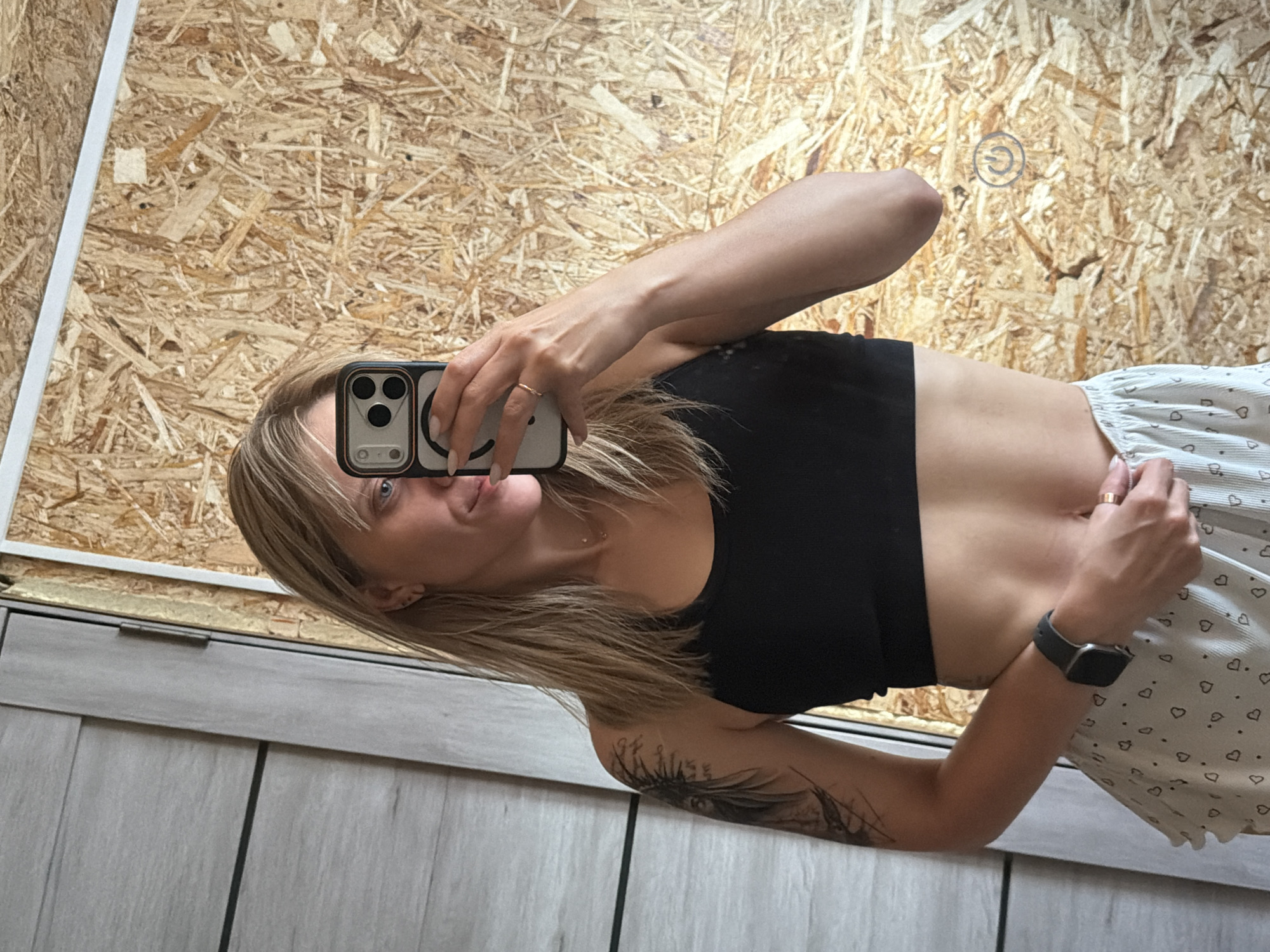 A woman with blonde hair takes a mirror selfie in what appears to be an unfinished interior space. She is wearing a black sports bra and patterned shorts. The background consists of a textured wall of compressed wood chips and a partially visible door. A power symbol is drawn on the wall. The woman is holding a smartphone to take the photo, and a tattoo is visible on her left arm. She is wearing a watch on her left wrist and a ring on her right index finger. The lighting suggests it is daytime. The location is an apartment in Chaikovsky, Russia.