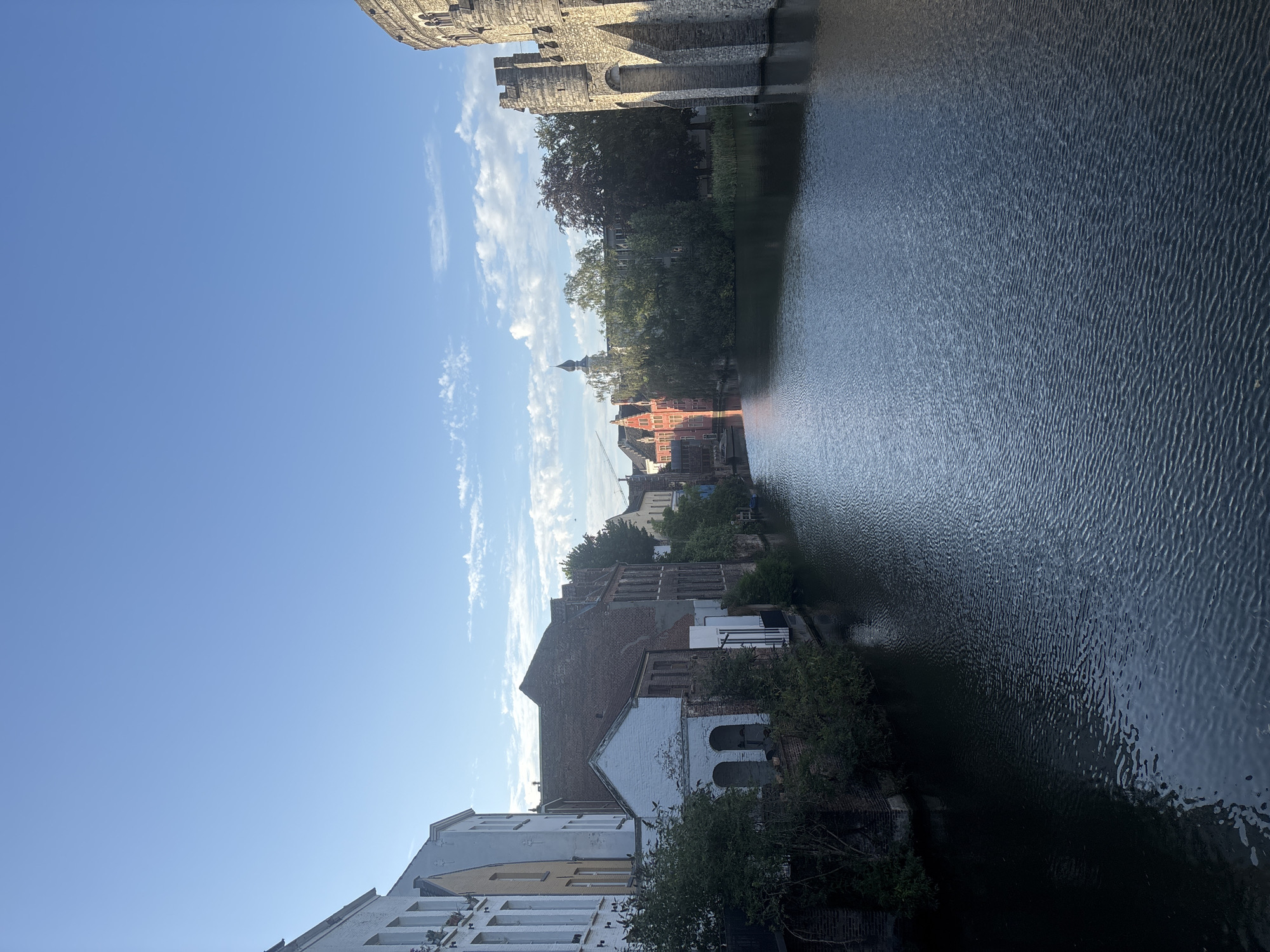 The image captures a picturesque canal scene in Ghent, Belgium, likely during the morning given the soft lighting. The water of the canal is calm with gentle ripples, reflecting the blue sky and surrounding architecture. On the left bank, a row of historic buildings with white and yellow facades lines the canal, some featuring arched windows and balconies. Lush green trees and bushes grow along the water's edge, softening the urban landscape.
Further down the canal, on the left, a striking cluster of red-brick buildings with characteristic Flemish architecture stands out. A spire-like tower is visible behind these buildings. To the right of the canal, a formidable stone structure with battlements, reminiscent of a medieval castle or fortress, dominates the view. The sky above is a clear, bright blue with a few wispy white clouds. No people are visible in the scene. The overall impression is one of historic charm and serene beauty.