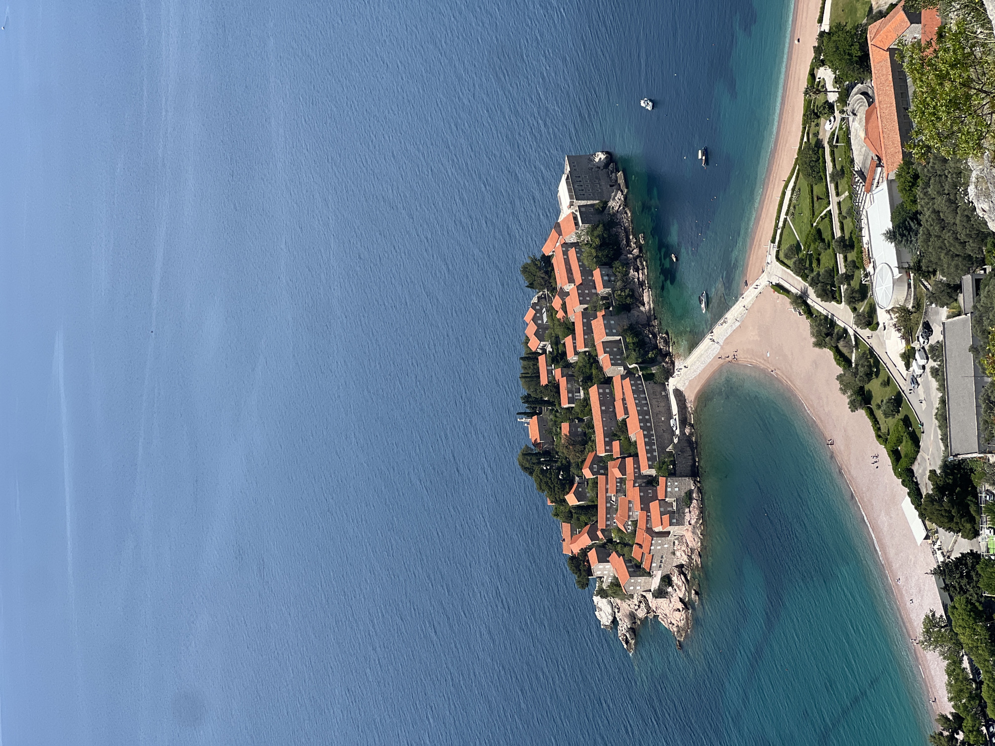 The image shows an aerial view of Sveti Stefan, Montenegro. A peninsula is densely populated with buildings featuring orange tile roofs. The peninsula connects to the mainland by a sandy isthmus, forming two beaches on either side. The water surrounding the area is a deep blue, contrasting with the lighter blue-green closer to the shore. On the mainland side, manicured gardens and building complexes are visible.