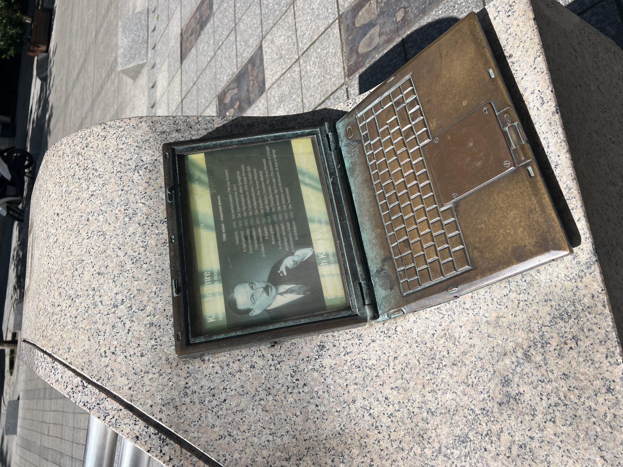 The image shows a bronze memorial resembling an open laptop, set into a granite structure, likely located on a street in Budapest, Hungary. The "screen" of the laptop displays a black and white photograph of Imre Kalman, along with a list of his important works in Hungarian and English. The "keyboard" side of the open laptop is also cast in bronze, depicting a detailed keyboard layout. The granite structure on which the memorial sits is mottled with black and gray specks. The surrounding environment consists of sidewalk paving stones and glimpses of vegetation and buildings. The lighting suggests a sunny day.
