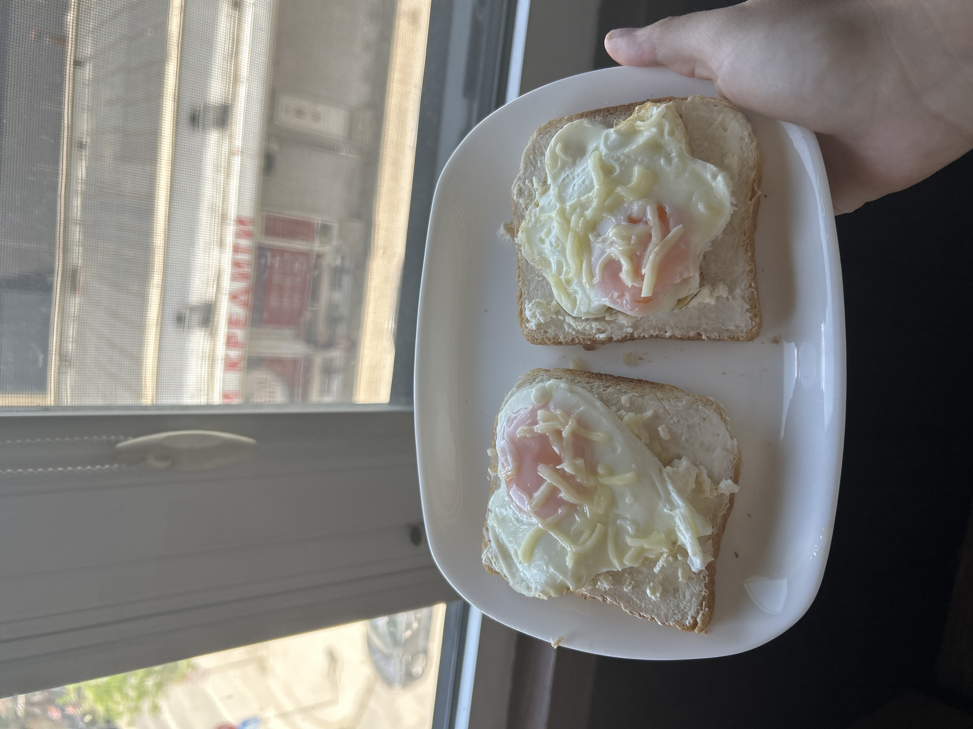 A light-skinned hand holds a white rectangular plate in the foreground, presenting two slices of toast each topped with a fried egg and a generous amount of shredded cheese or a creamy white spread. The eggs display a soft, pinkish yolk. The background reveals an outdoor urban environment viewed through a window with a visible screen grid. To the left, a blurred street scene includes a green tree and a dark-colored vehicle. To the right, an adjacent building features a prominent red sign with Cyrillic text, clearly displaying "КРЕАМИ" (KREAMI), positioned over smaller red rectangular elements. This Cyrillic signage identifies the location as the city of Sofia, Bulgaria. A white, oval-shaped cord pull for window blinds is visible hanging from the left window frame. The scene is illuminated by natural daylight.